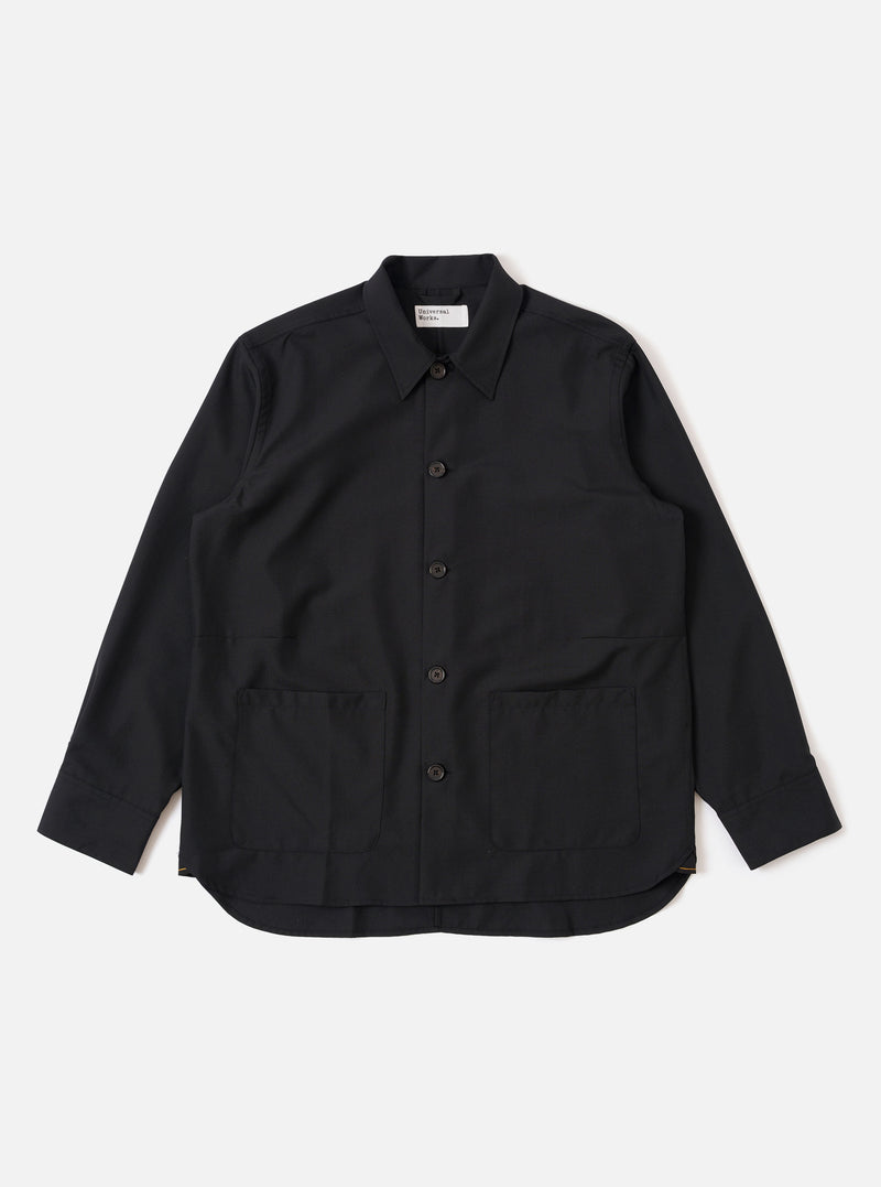 The Black Tropical Suiting Travail Overshirt is shown as a flat lay against a white background. The overshirt features a one-piece collar, a central button-down closure, and two large patch pockets at the waist. The lightweight black tropical suiting fabric has a smooth, minimal appearance.
