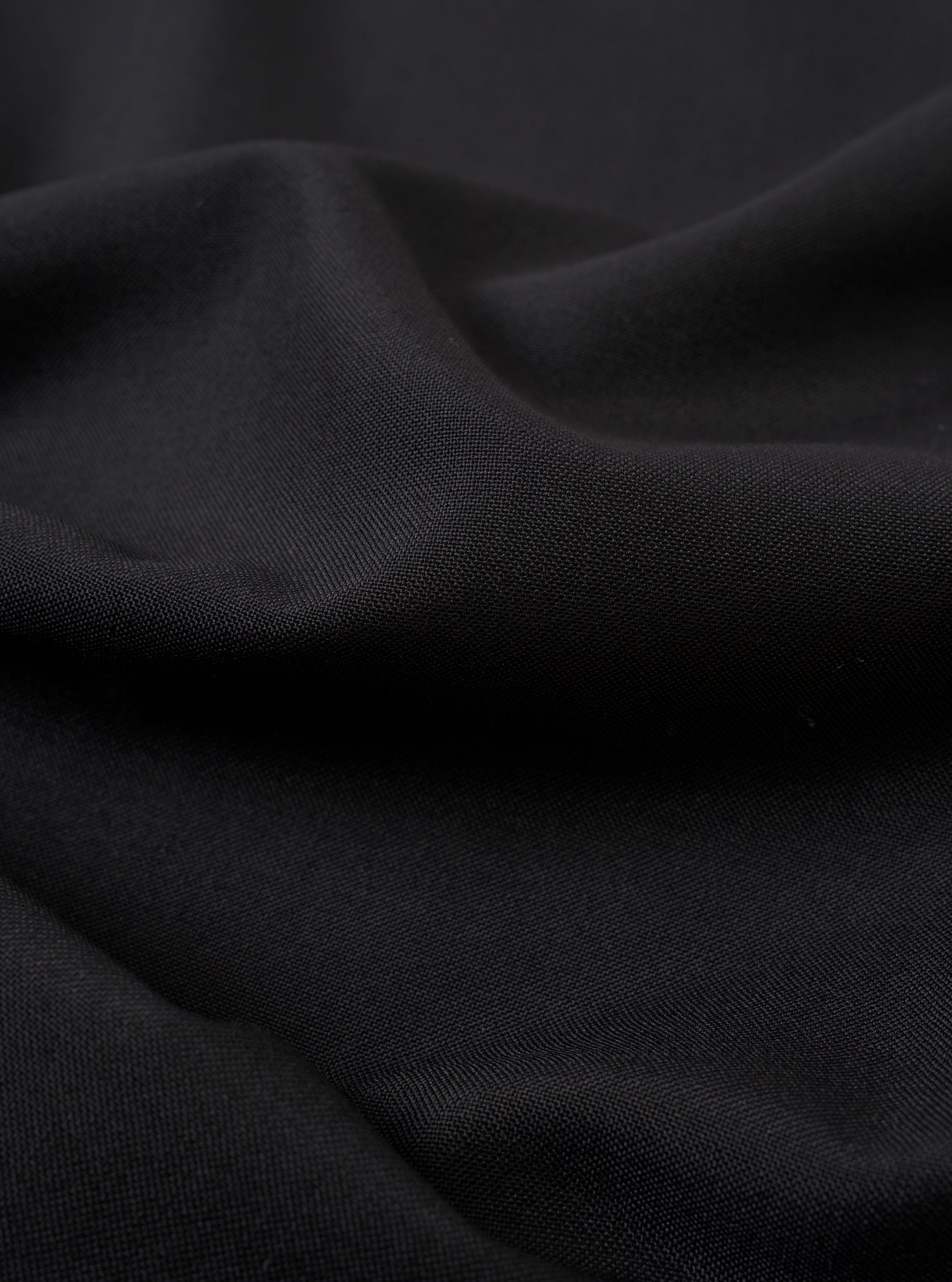 A close-up swatch image of the Black Tropical Suiting Travail Overshirt fabric. The image focuses on the textile detail, showing a solid black color with a smooth, lightweight weave characteristic of tropical suiting. The material fills the entire frame, highlighting its clean and professional finish.