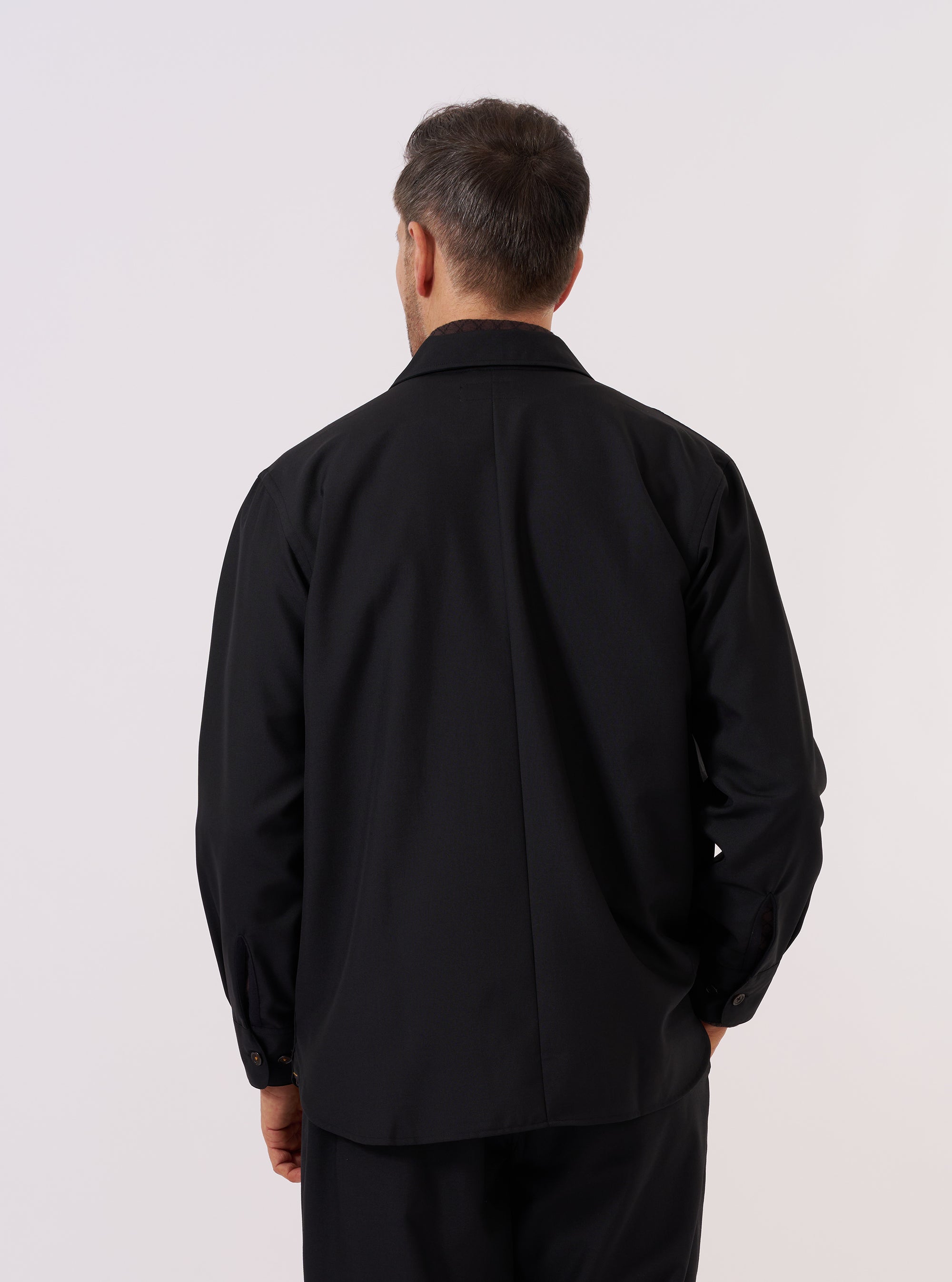 A model wears the Black Tropical Suiting Travail Overshirt, shown from the back against a plain white background. The overshirt features a structured collar, a straight hem, and a relaxed fit. The garment is constructed from a smooth black tropical suiting fabric with a lightweight, matte finish.