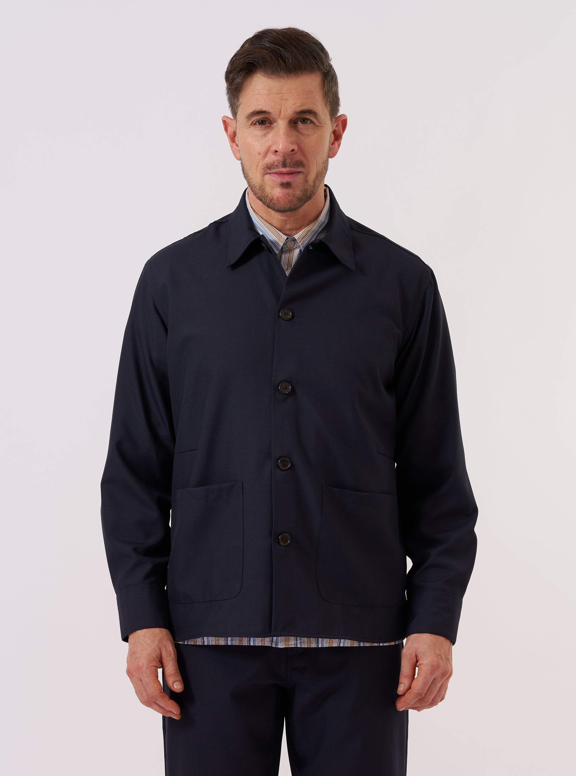 A model wears the Navy Tropical Suiting Travail Overshirt over a light-colored top, standing against a plain white background. The overshirt features a loose, unstructured fit, a one-piece collar, a central button-down front, and two large patch pockets at the waist. The navy blue tropical suiting fabric appears lightweight and smooth.