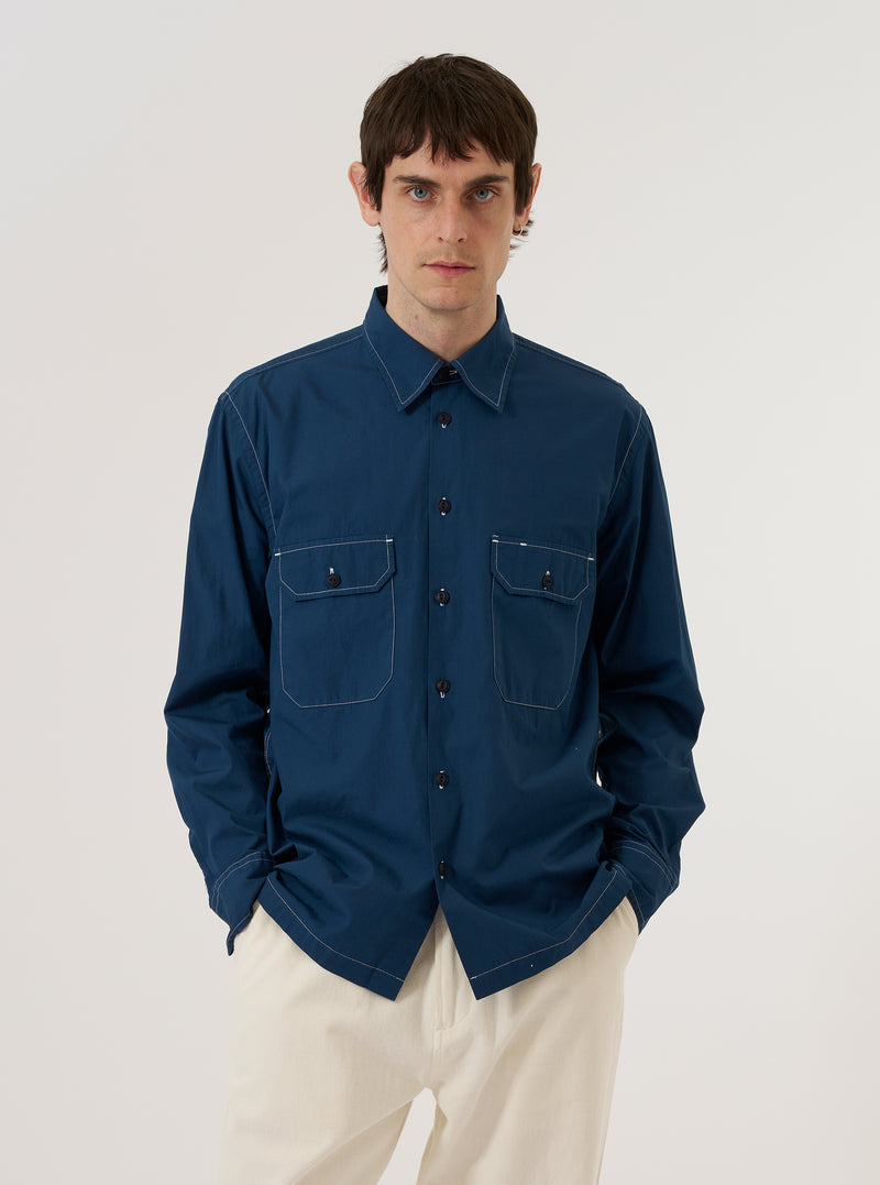 A model wears the Navy Organic Fine Poplin CPO Shirt against a plain, neutral studio background. The shirt features a button-down front, a classic pointed collar, and two prominent chest patch pockets with button-flap closures. The navy organic cotton poplin fabric has a smooth, fine-grained finish. The model is shown in a medium shot, wearing the shirt as an outer layer.