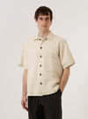 A model wears the Ecru Koko Cotlin S/S Fatigue Shirt standing against a plain white background. The shirt features a central button-down closure, a classic collar, and two prominent patch pockets on the chest. The short-sleeved garment is made from a light ecru fabric with a textured cotton-linen finish.