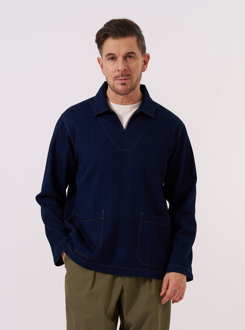 A model wears the Indigo Recycled Herringbone Smock Overshirt against a plain, neutral studio background. The overshirt features a pullover smock design with a buttoned half-placket, a structured collar, and three large patch pockets on the front. The dark indigo fabric displays a distinct herringbone weave texture throughout.