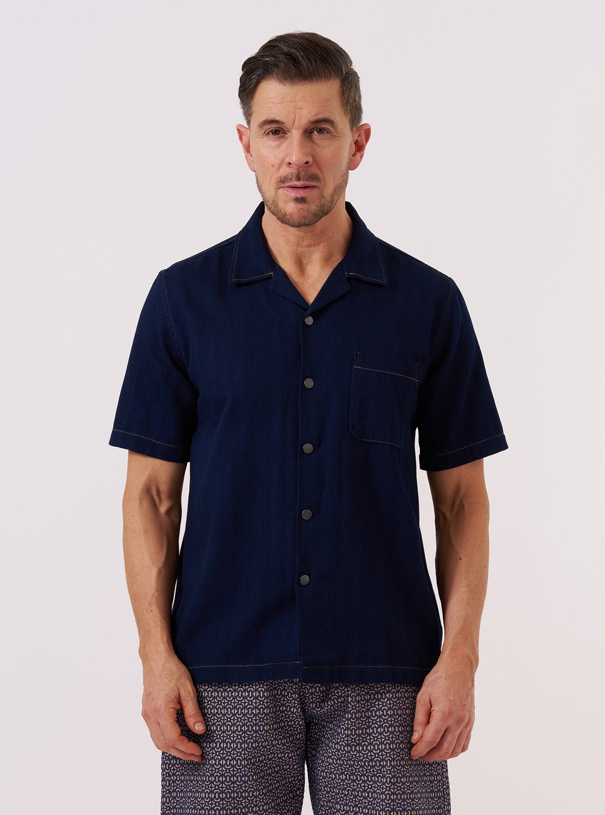 A model wears the Indigo Recycled Herringbone Road Shirt, standing against a plain, neutral background. The shirt features a relaxed camp collar, a button-down front, and a single patch pocket on the chest. The indigo blue fabric displays a visible herringbone weave texture throughout.