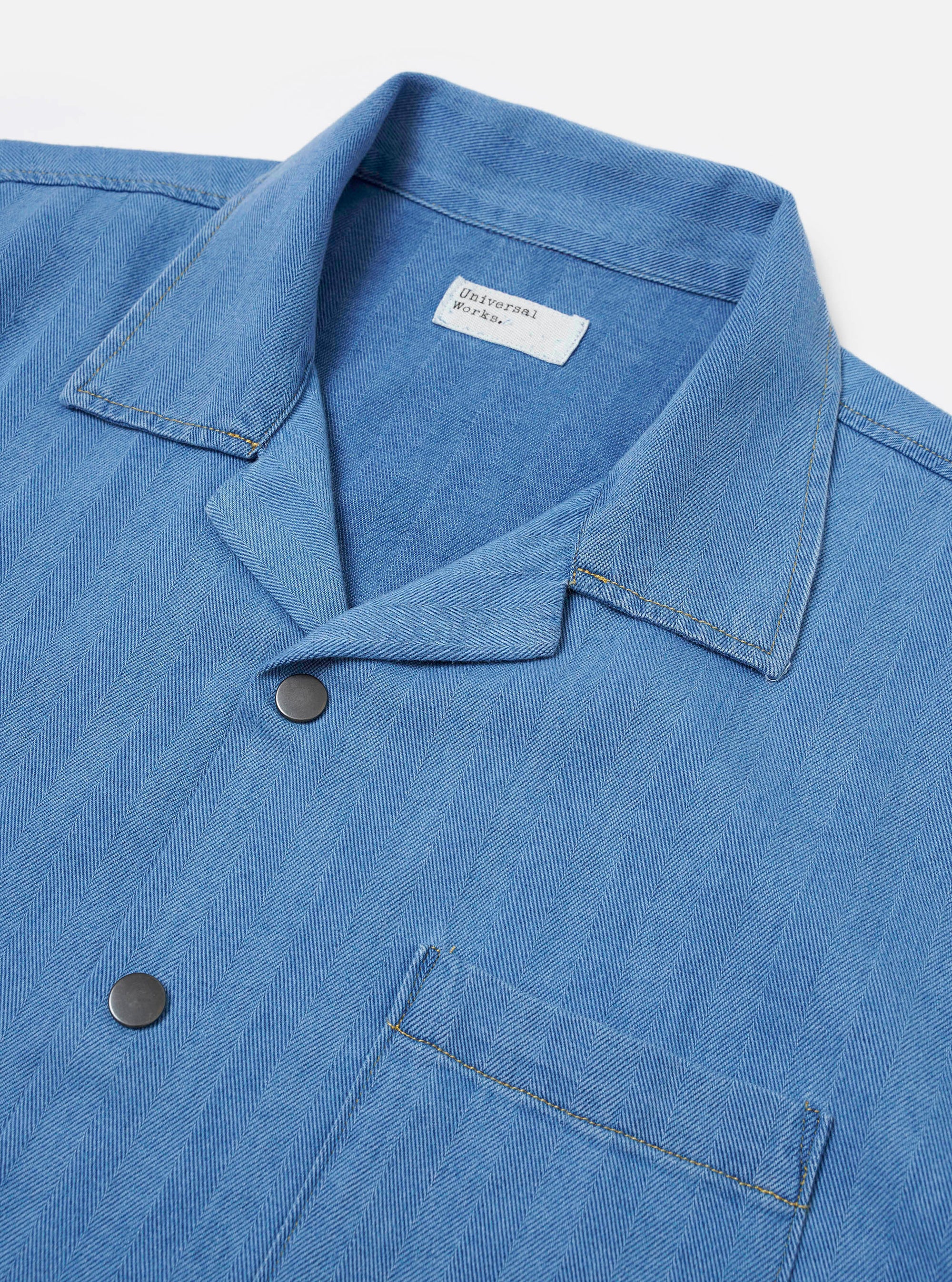 The Washed Indigo Recycled Herringbone Road Shirt is shown from a rear angle as a flat lay against a plain white background. The image highlights the shirt's straight hem, simple back yoke, and the distinct V-shaped pattern of the indigo-dyed herringbone weave.