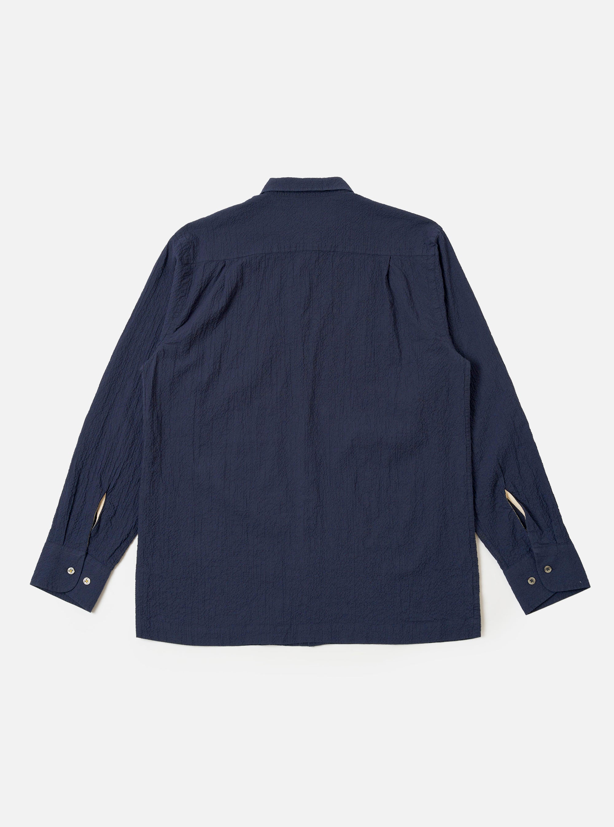 The Navy Seersucker Shirting Lucky Shirt is shown from the back as a flat lay against a plain white background. The image highlights the shirt's relaxed fit, straight hem, and the subtle puckered texture of the navy blue seersucker fabric. A simple back yoke and clean stitching along the hem and sleeves are visible.
