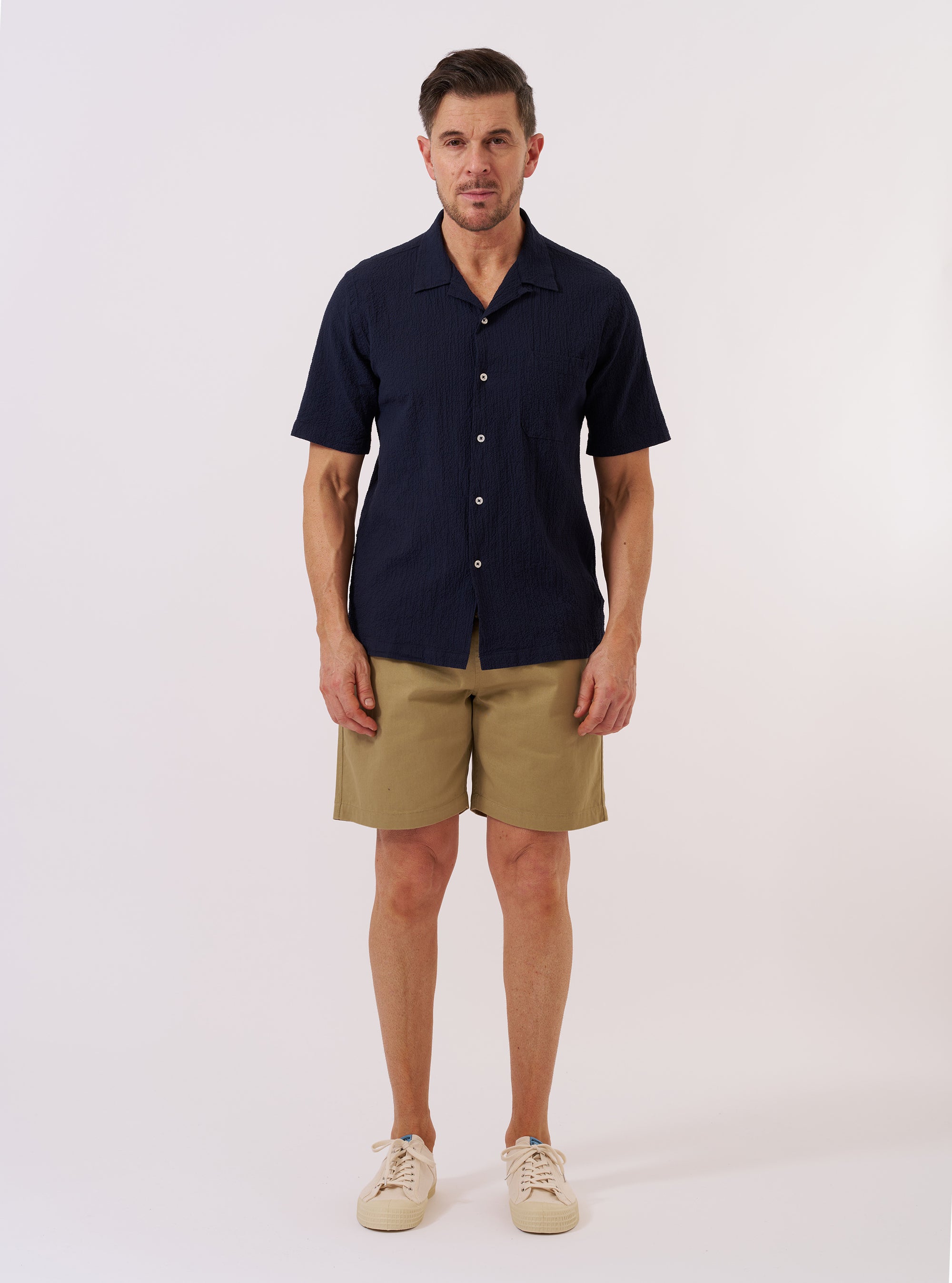The Navy Seersucker Shirting Road Shirt is shown as a flat lay against a plain white background. The shirt features a camp collar, a central button-down front with tonal buttons, and a single chest patch pocket on the left side. The fabric has a distinct puckered seersucker texture in a deep navy blue.