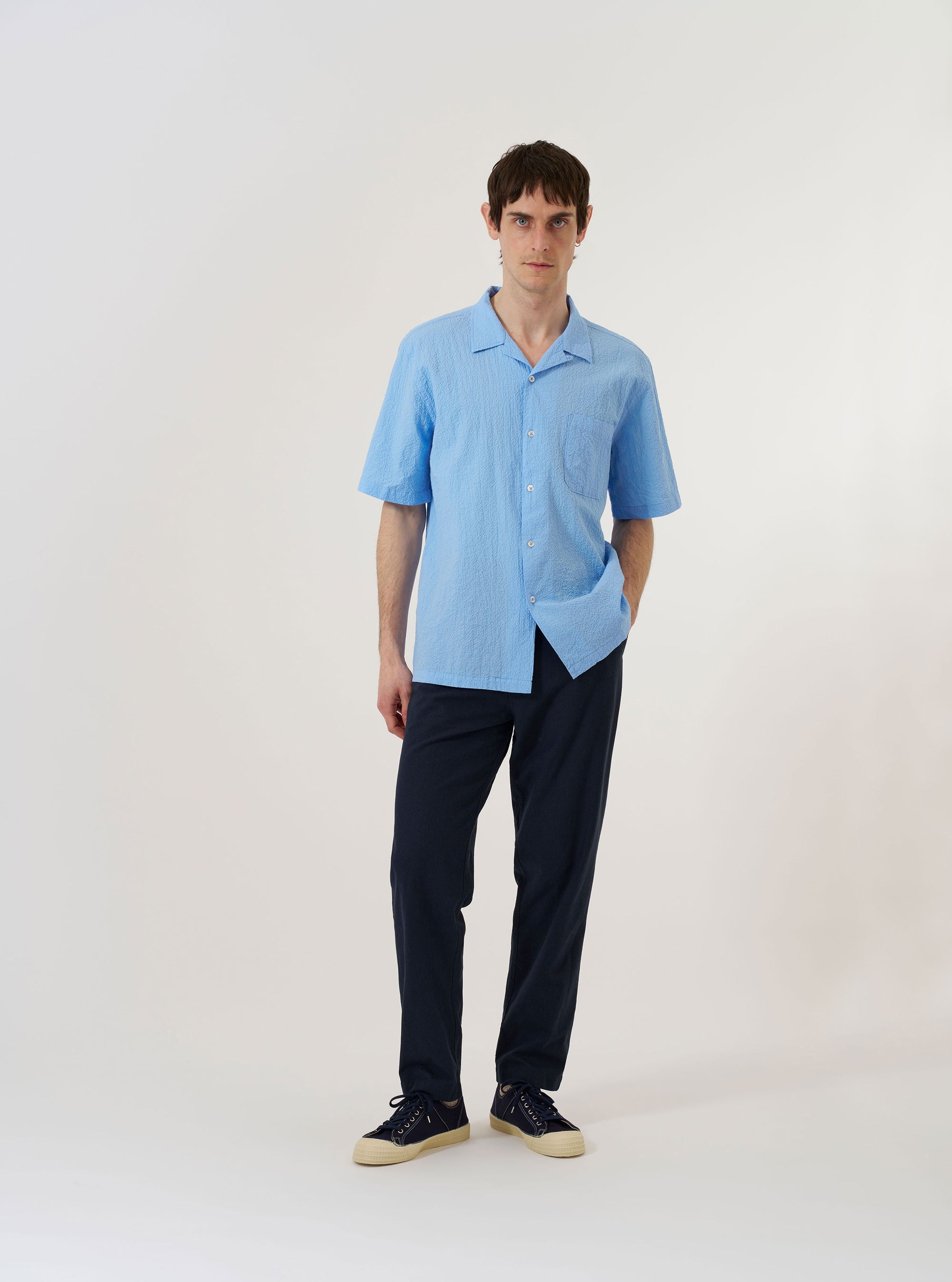 The Sky Blue Seersucker Shirting Road Shirt is shown as a flat lay against a plain white background. The shirt features a camp collar, short sleeves, and a central button-down front with tonal buttons. A single patch pocket is located on the left chest. The fabric has a distinct puckered seersucker texture in a light sky blue color.
