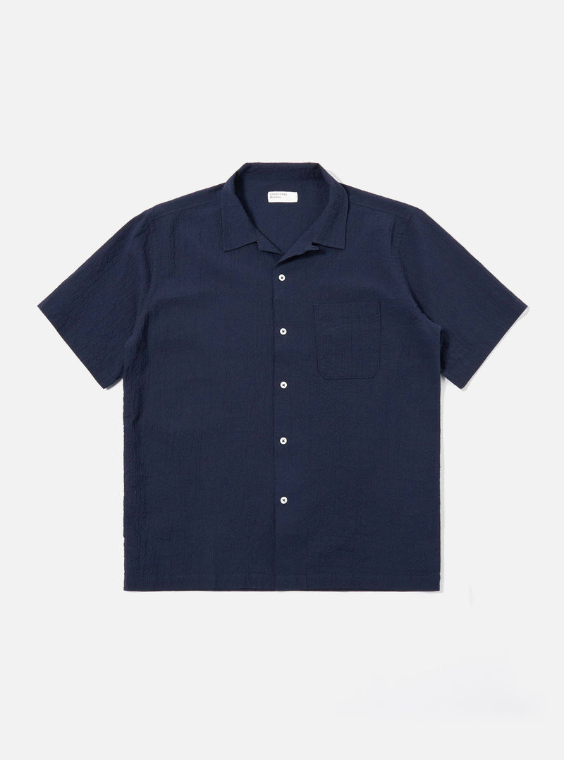 The Navy Seersucker Shirting Road Shirt is shown as a flat lay against a plain white background. The shirt features a camp collar, a central button-down front with tonal buttons, a single chest patch pocket, and short sleeves. The dark navy fabric has a distinct puckered seersucker texture.