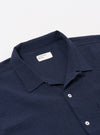The Navy Seersucker Shirting Road Shirt is shown from the back as a flat lay against a plain white background. The image highlights the shirt's straight hem, short sleeves, and the distinct puckered texture of the navy blue seersucker fabric. A horizontal yoke across the shoulders and a simple, clean construction are visible.