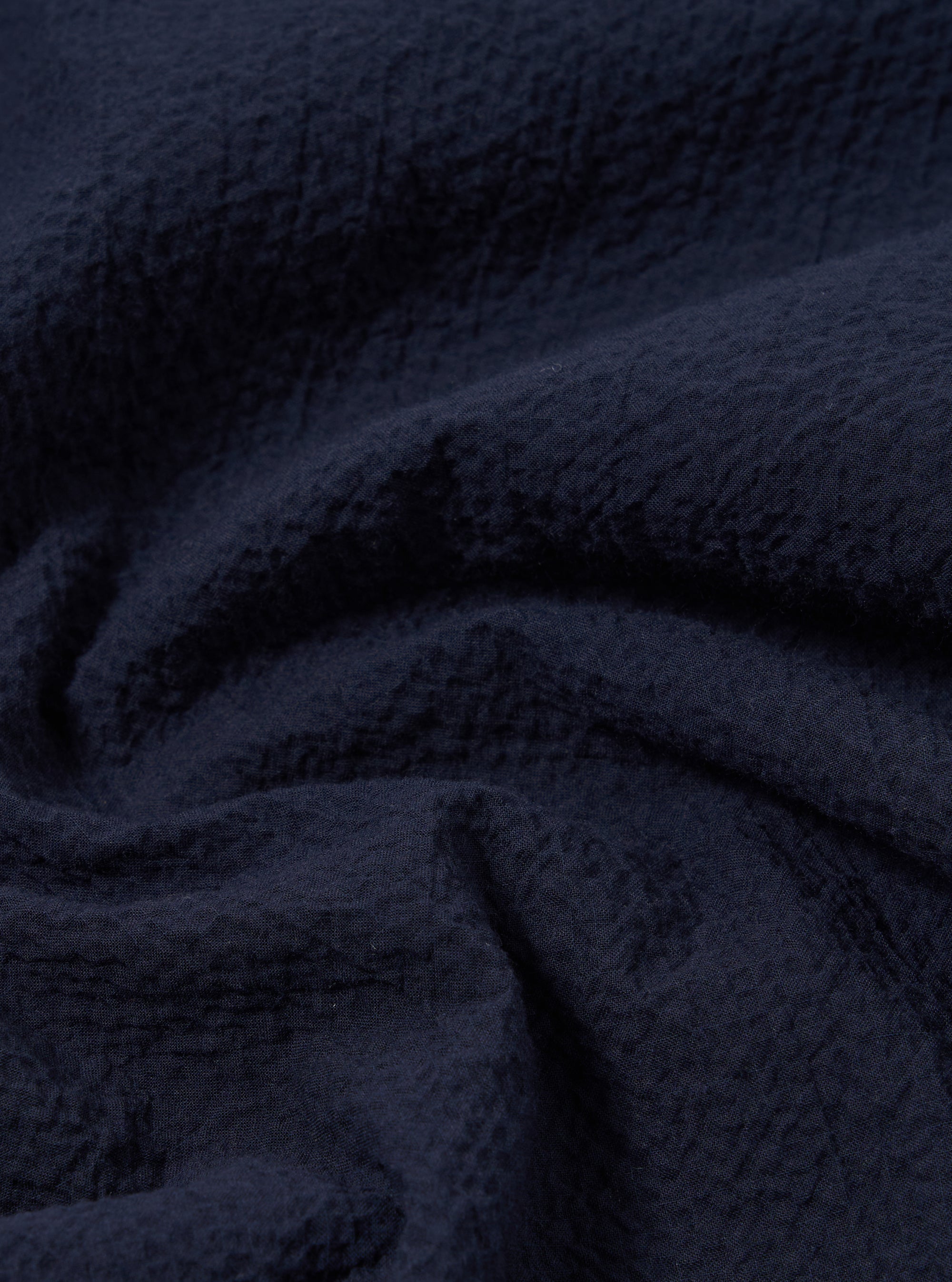 A close-up swatch image of the Navy Seersucker Shirting Road Shirt fabric highlights the deep navy blue color and the distinct puckered, wavy texture characteristic of seersucker. The material fills the frame, providing a detailed view of the lightweight cotton fabric's weave.