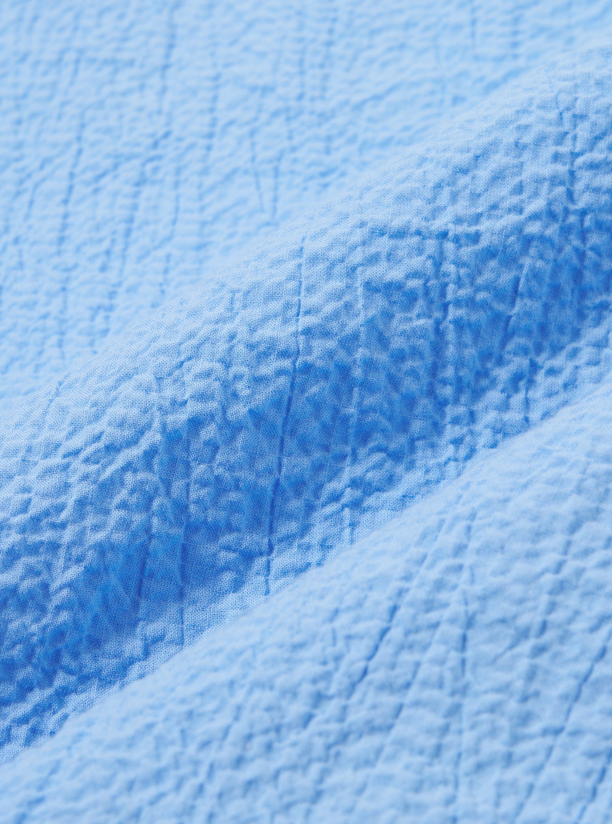 A close-up swatch image of the Sky Blue Seersucker Shirting Road Shirt fabric highlights the light blue and white striped pattern and the characteristic puckered texture of the seersucker weave. The fabric fills the frame, providing a detailed view of the material's construction.