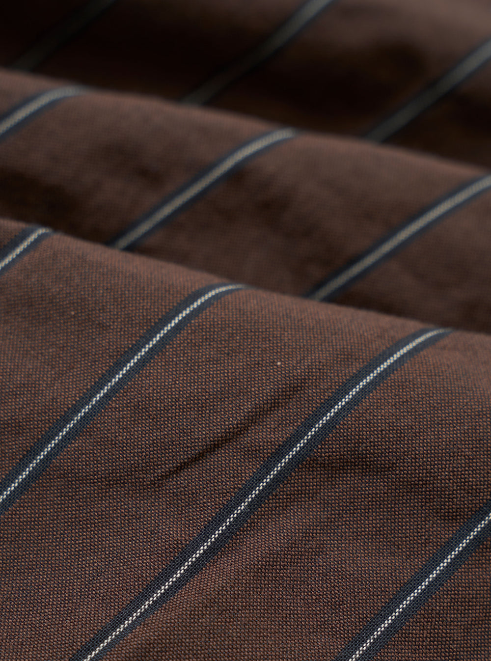 A close-up fabric swatch of the Brown Chester Stripe Oxford Pant highlights the texture and pattern of the material. The image displays a brown base with thin, light-colored vertical stripes across the cotton fabric.