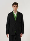 A model wears the Black Valencia Cotton Two Button Jacket over a light-colored shirt, standing against a plain, neutral background. The jacket features a classic two-button front closure, notched lapels, and three front patch pockets. The black fabric has a subtle, textured appearance.