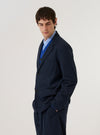 A model wears the Navy Valencia Cotton Two Button Jacket over a light-colored shirt, shown from a side angle against a plain, neutral studio background. The jacket features a classic two-button front closure, notched lapels, and functional patch pockets at the waist. The navy fabric has a soft, structured appearance consistent with a cotton-blend suiting material.