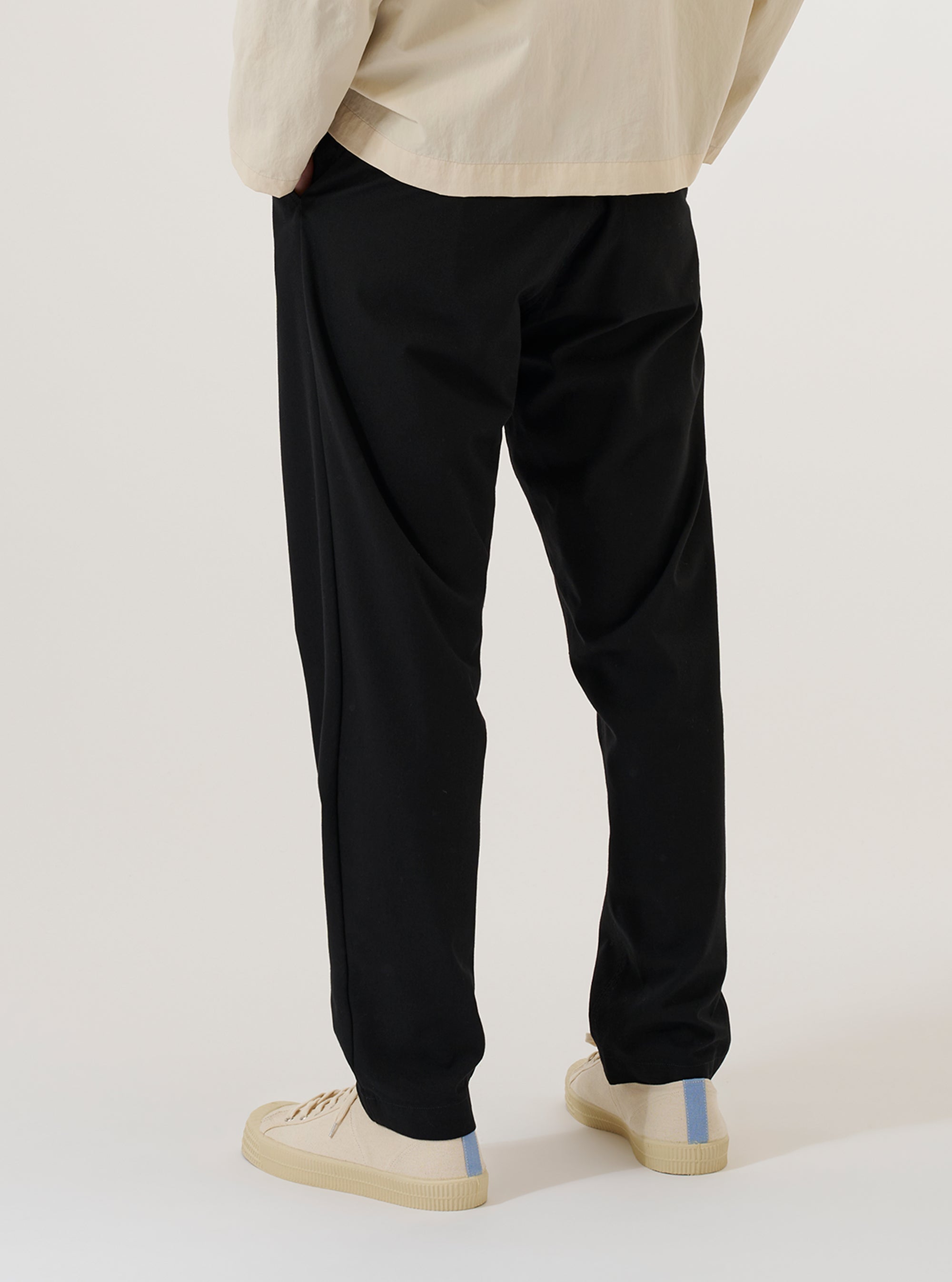 A model wears the Black Valencia Cotton Military Chino, shown from a side profile against a plain white background. The image highlights the straight-leg fit and the clean construction of the black Valencia cotton fabric. The model is wearing the trousers from the waist down, paired with a light-colored top and dark shoes.