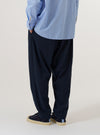 A model is wearing the Navy Valencia Cotton Pleated Track Pant, shown from a rear three-quarter angle against a plain white background. The image highlights the loose, tapered fit of the trousers and the clean finish of the navy Valencia cotton fabric. One rear buttoned flap pocket is visible, and the model is wearing the pants with brown shoes and white socks.