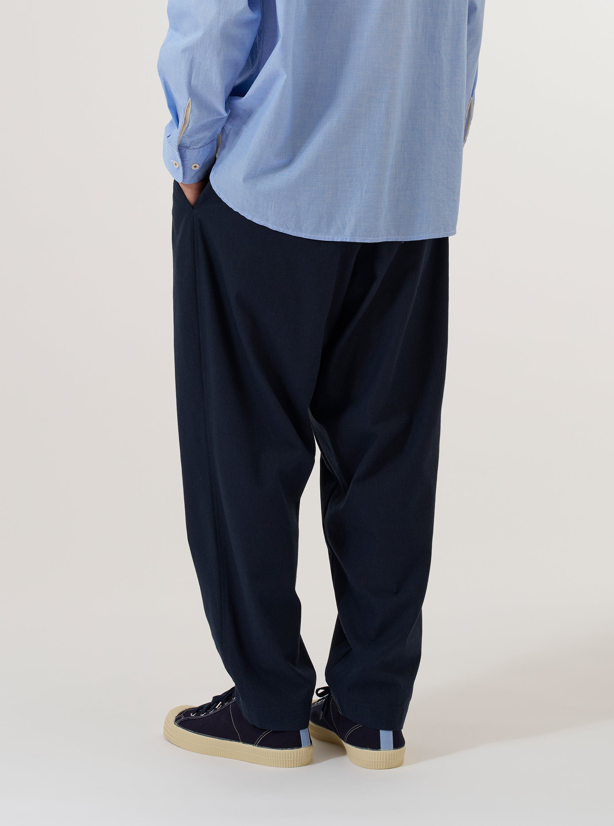 A model is wearing the Navy Valencia Cotton Pleated Track Pant, shown from a rear three-quarter angle against a plain white background. The image highlights the loose, tapered fit of the trousers and the clean finish of the navy Valencia cotton fabric. One rear buttoned flap pocket is visible, and the model is wearing the pants with brown shoes and white socks.
