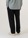 A model wears the Black Valencia Cotton Pleated Track Pant, shown from the waist down against a plain white background. The image highlights the loose, tapered fit of the trousers, the single pleat at the front, and the elasticated waistband with a drawstring. The black cotton fabric has a smooth finish, and the model is wearing the pants with a white t-shirt and white socks.