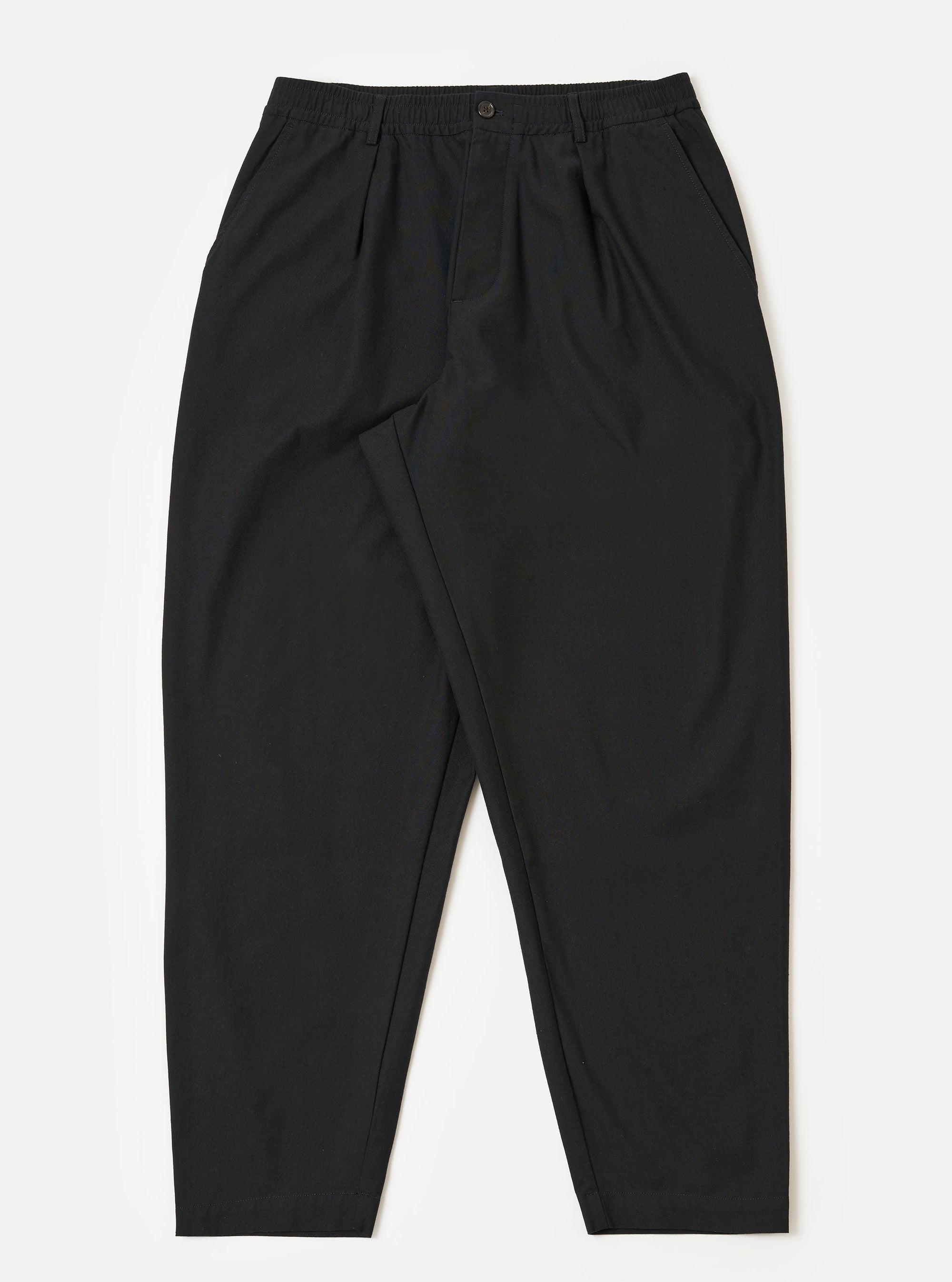 The Black Valencia Cotton Pleated Track Pant is shown as a flat lay against a white background. The trousers feature a loose, tapered leg with a single pleat at the front and an elasticated drawstring waistband. The fabric is a smooth black cotton blend.