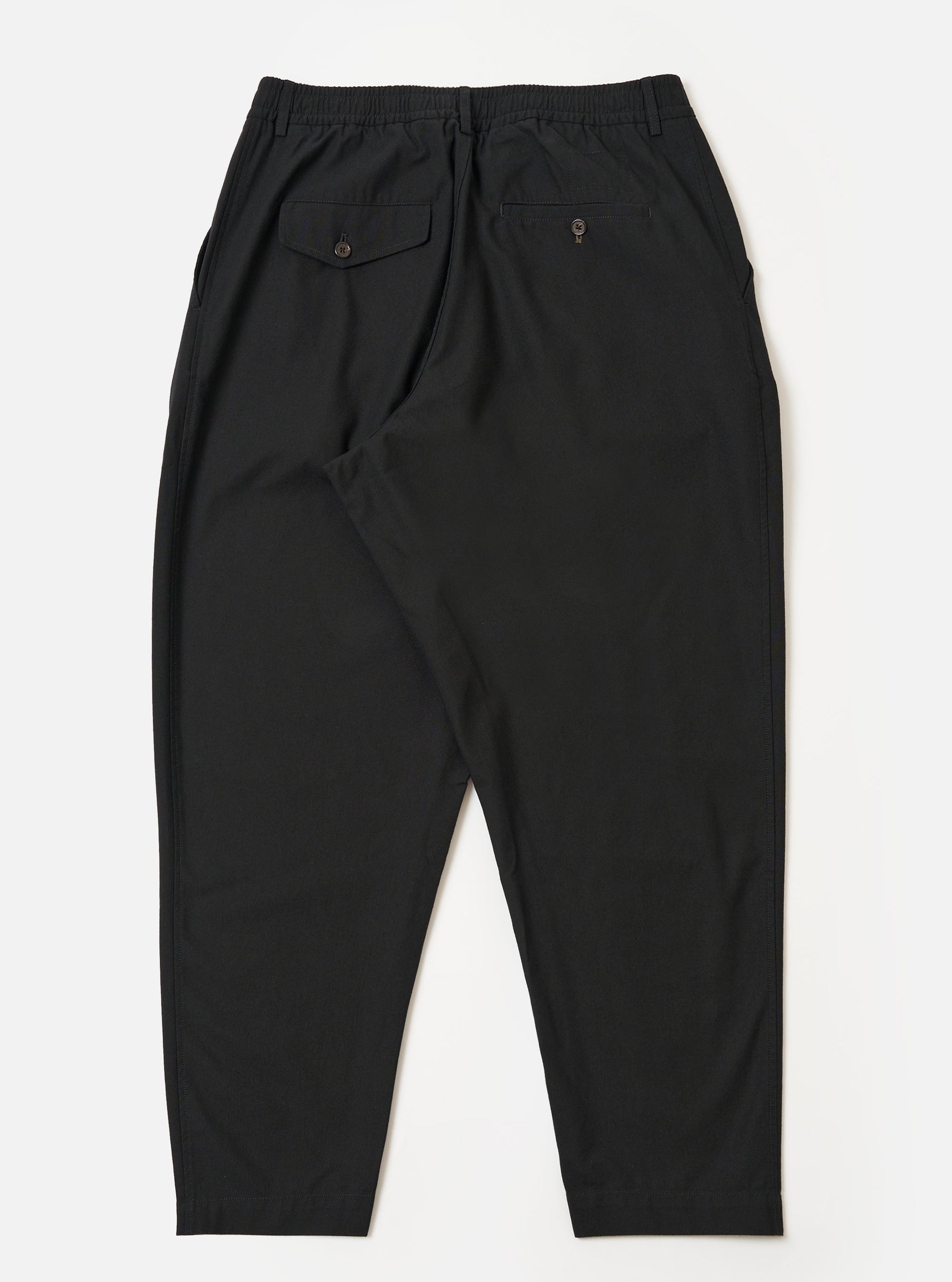 The Black Valencia Cotton Pleated Track Pant is shown from the back as a flat lay against a plain white background. The image highlights the elasticated waistband and the two rear pockets: one featuring a buttoned flap and the other a button jetted design. The black fabric has a clean, fine plain weave texture.