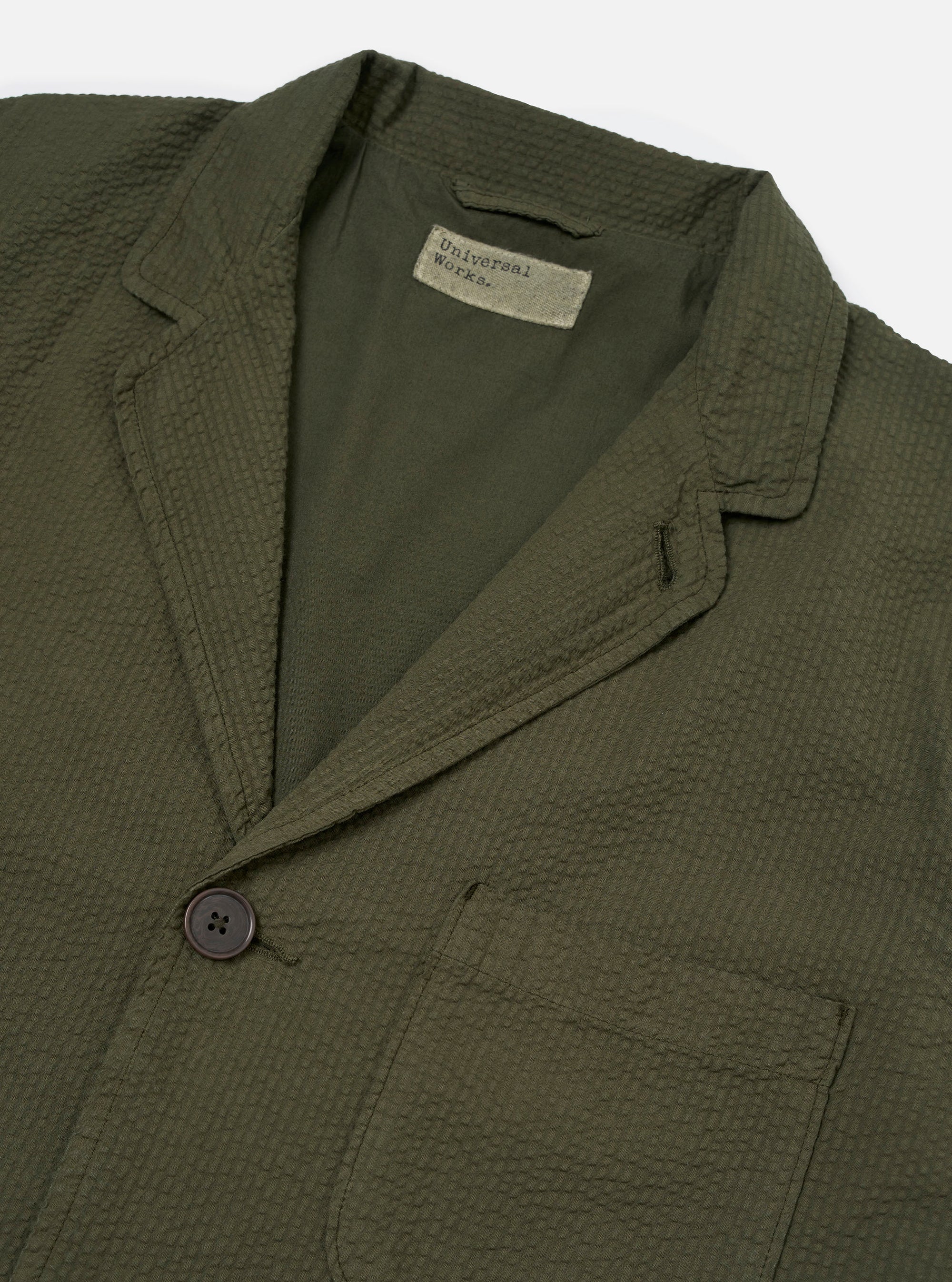The Olive Merida Seersucker Three Button Jacket is shown as a flat lay against a white background. The jacket features a three-button front closure, a notch lapel, and two large patch pockets at the hips. The olive green fabric has a distinct puckered seersucker texture throughout.