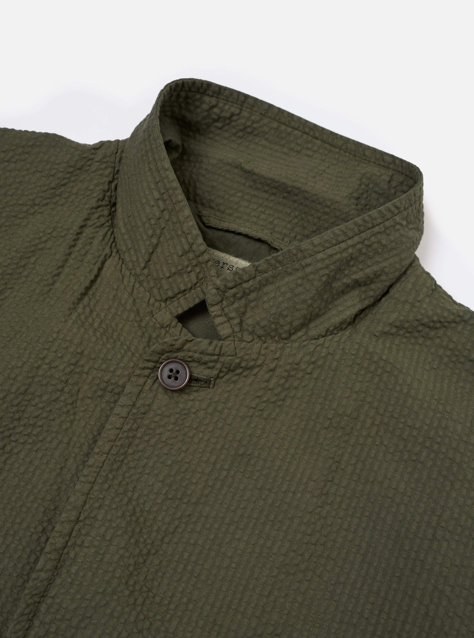 The Olive Merida Seersucker Three Button Jacket is shown as a flat lay against a white background. The jacket features three buttons, a classic lapel, one chest patch pocket, and two lower patch pockets. The olive green cotton fabric has a characteristic puckered seersucker texture.