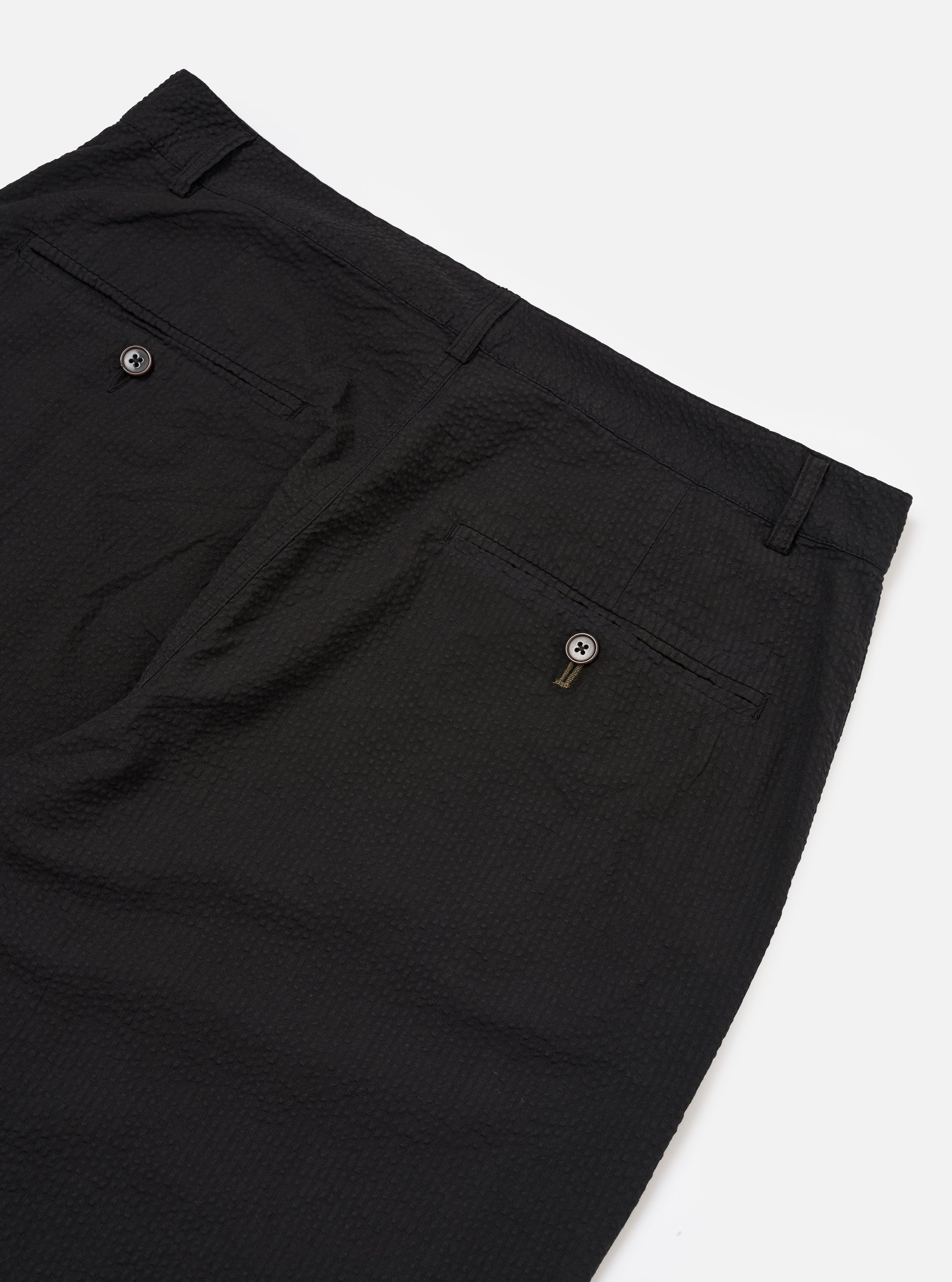 The Black Merida Seersucker Military Chino is shown from a rear angle as a flat lay against a plain white background. The image highlights the two buttoned flap back pockets and the "Hollywood" one-piece waistband. The black cotton fabric features a distinct puckered seersucker texture throughout.