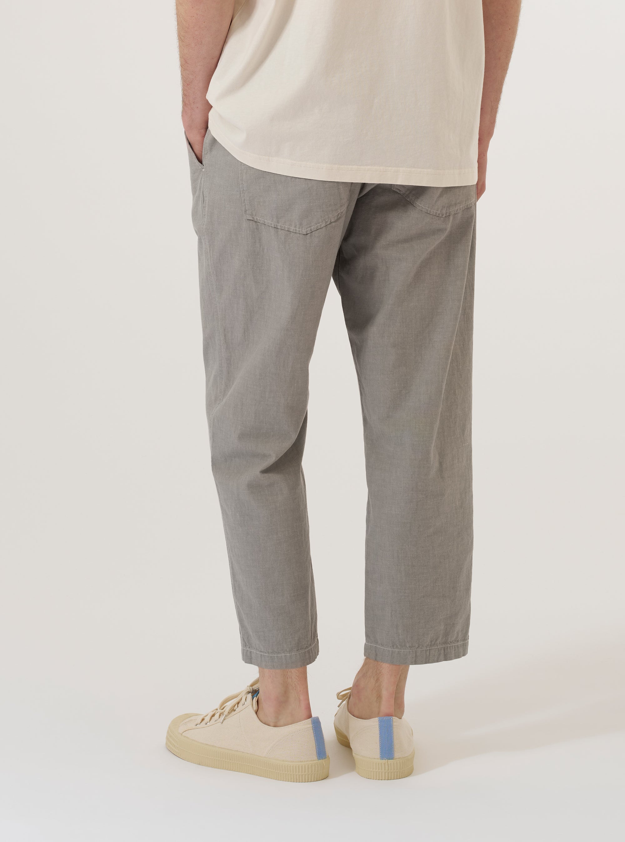 A model wears the Grey Izumi Cotton Hi Water Trouser, shown from the waist down from a rear angle against a plain white background. The trousers feature a slim, cropped fit, an elasticated waistband with a drawstring, and two large rear patch pockets. The grey cotton fabric has a visible textured appearance.