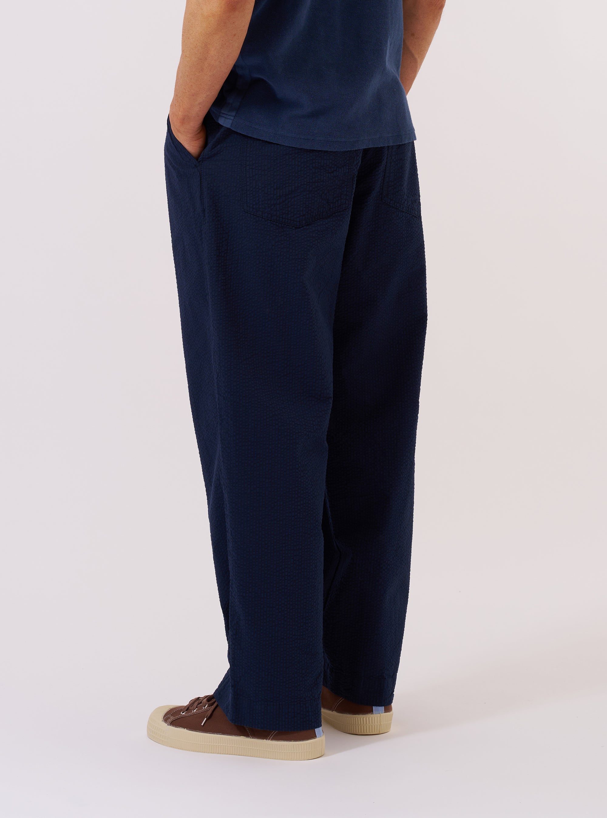 A model wears the Navy Derby Stripe Duke Pant against a plain, neutral background. The trousers are shown from a rear angle, highlighting the loose, wide-leg fit and the vertical navy stripe pattern. The garment features a relaxed silhouette with a structured waistband and a straight hem.