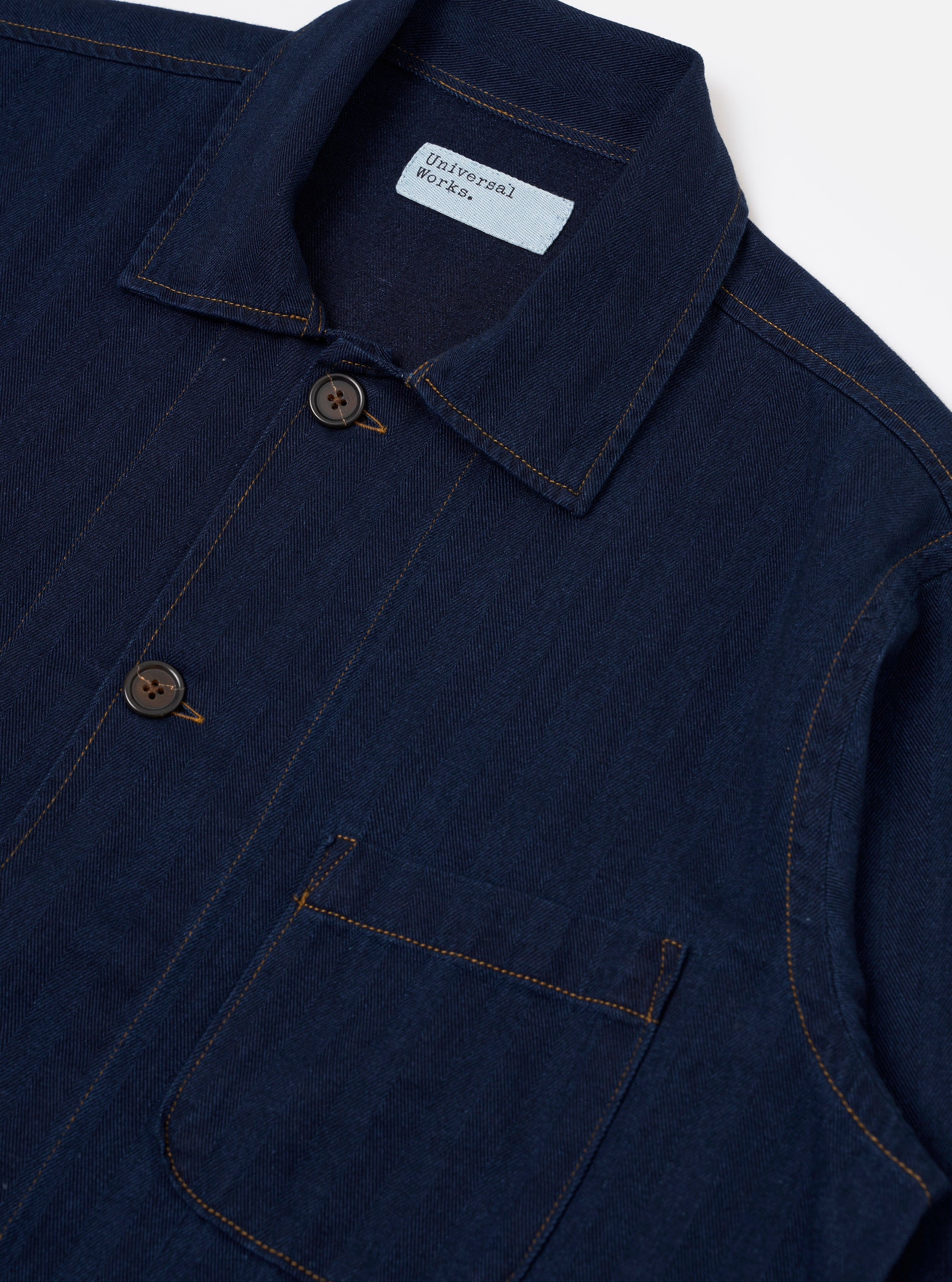 The Indigo Recycled Herringbone Bakers Overshirt is shown from the back as a flat lay against a plain white background. The image highlights the straight hem, the simple one-piece back panel, and the textured indigo blue herringbone weave of the recycled cotton fabric.