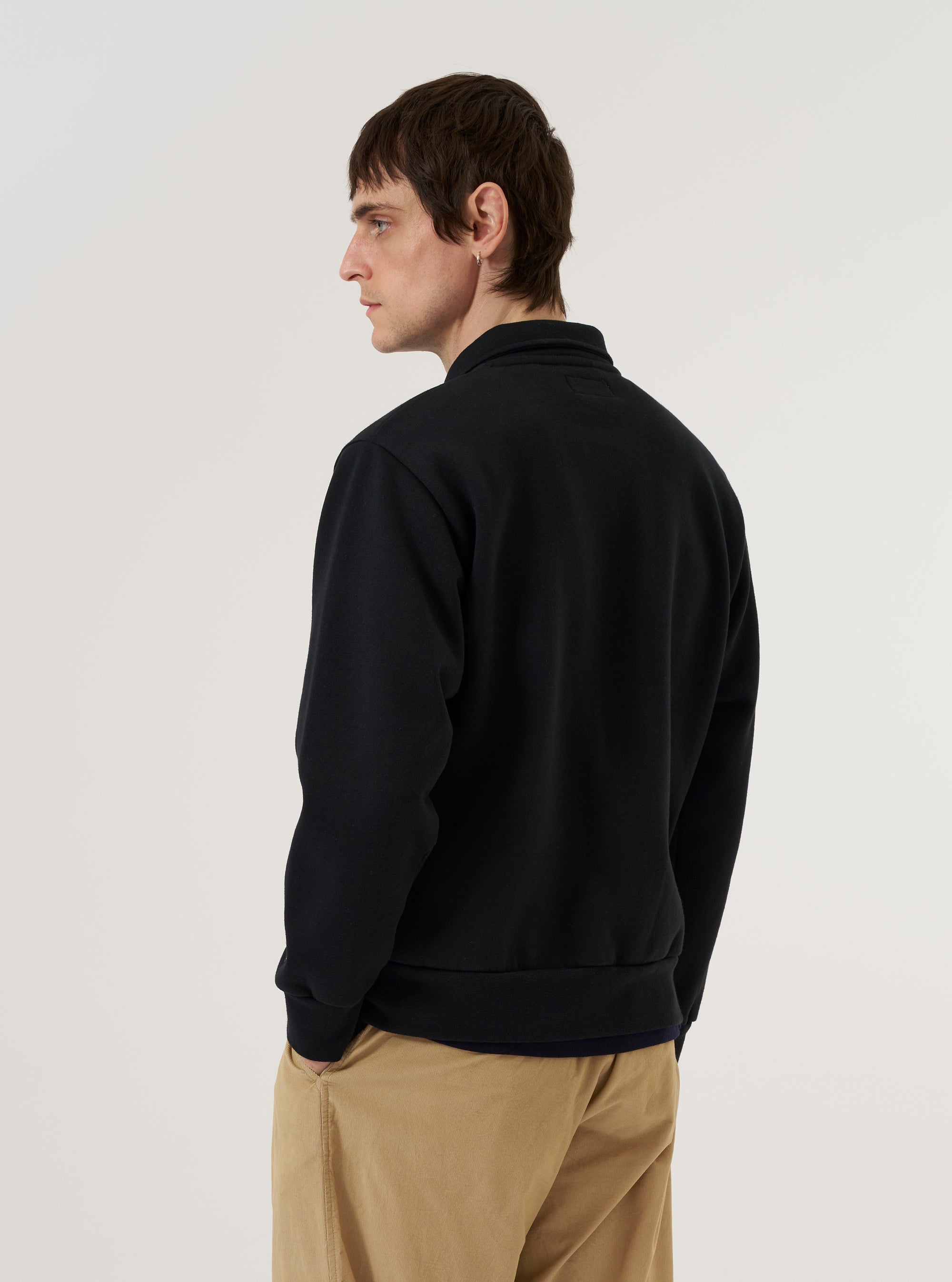 A model wears the Black Recycled Cotton Blend Jersey Tyler Jacket, shown from a rear three-quarter angle against a plain white background. The jacket features a relaxed fit with ribbed cuffs and a ribbed hem. The smooth, black jersey fabric and the self-fabric collar are prominent. The model is wearing the jacket over a light-colored top and dark trousers.
