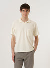 A model wears the Ecru Piquet Vacation Polo against a plain white background. The polo features a buttonless open neck, a single-piece collar, and a single patch pocket on the left chest. The ecru-colored cotton piquet fabric has a visible fine honeycomb texture. The model is shown from the chest up, highlighting the relaxed fit and clean design of the neckline.