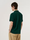 A model wears the Green Piquet Vacation Polo, which features a textured piqué cotton fabric, a single-piece collar, and a single patch chest pocket. The polo has a straight hem with small side vents and a slightly longer back hem. The model is shown from the chest down against a plain white background.