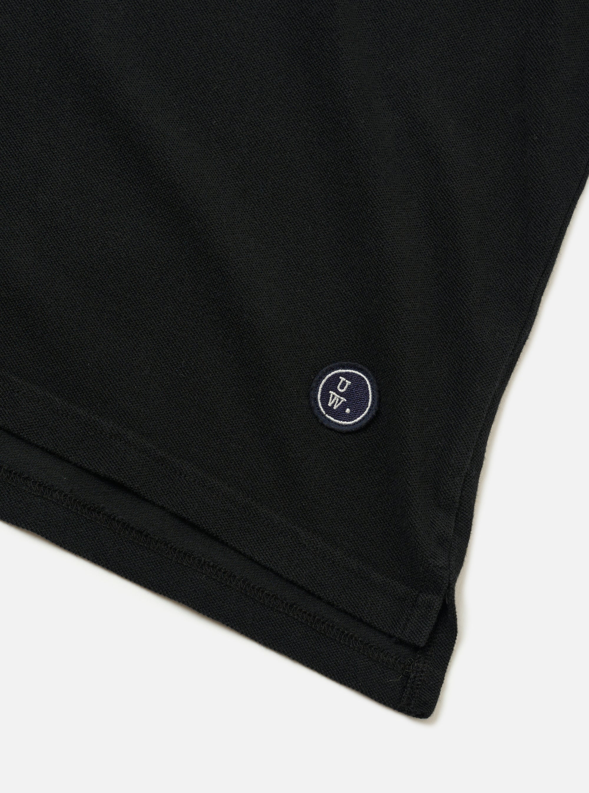 The Black Piquet Vacation Polo is shown as a flat lay against a plain white background. The image provides a close-up view of the back of the garment, highlighting the textured black piquet cotton fabric, a simple straight hem, and a one-piece collar.