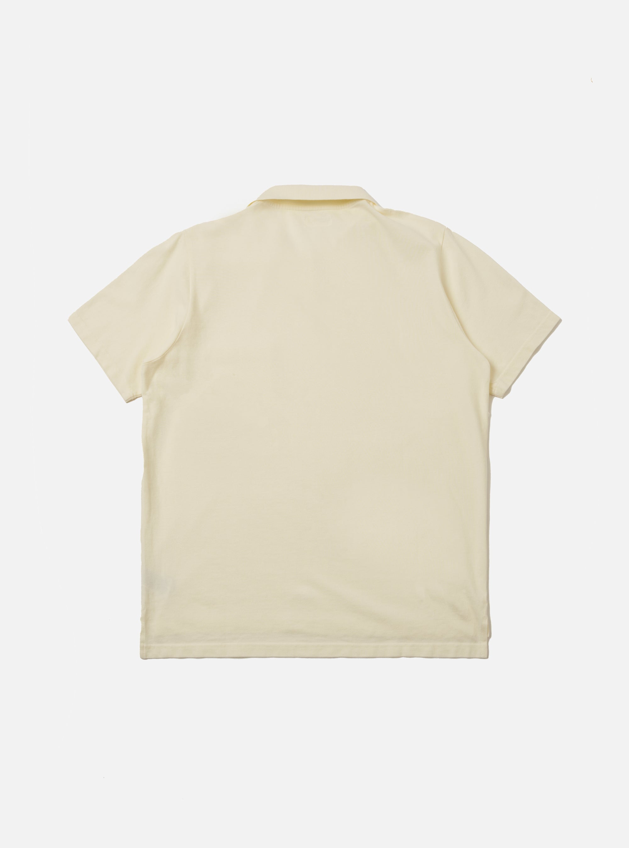 The Ecru Piquet Vacation Polo is shown from the back as a flat lay against a plain white background. The image highlights the clean construction of the garment's rear, the simple one-piece collar, and a dropped back hem with small side vents. The ecru-colored cotton fabric features a visible honey-comb piquet texture.