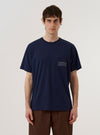 A model wears the Bright Navy Organic Jersey Print Pocket Tee against a plain, light-colored background. The t-shirt features a crew neckline, short sleeves, and a patch pocket on the left chest with a white graphic print. The garment is a vibrant navy blue and is shown from the front in a medium shot.