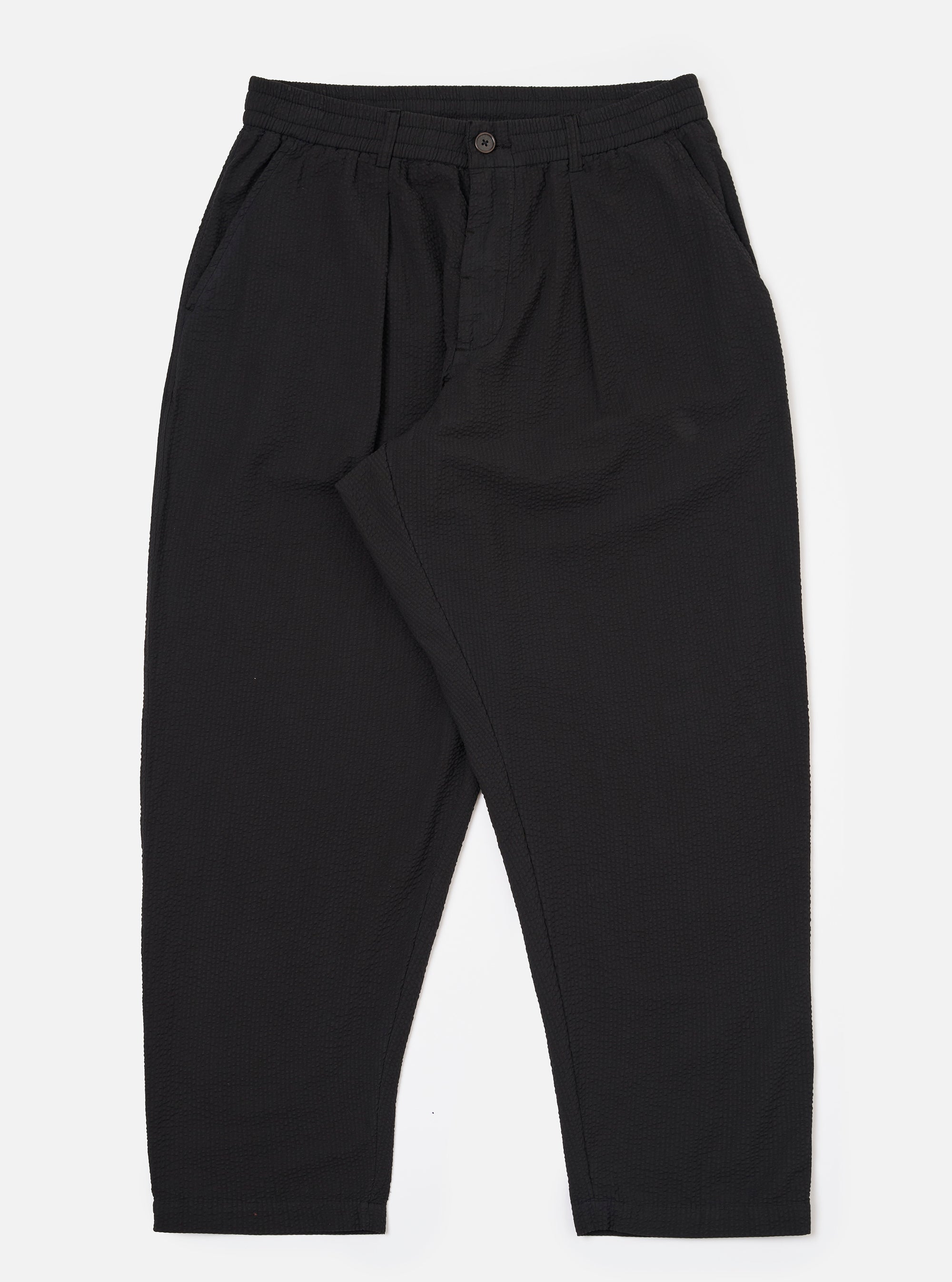 The Black Merida Seersucker Pleated Track Pant is shown as a flat lay against a white background. The trousers feature an elasticated waistband with a central drawstring, a single pleat on each leg, and two side pockets. The black cotton fabric has a distinct puckered seersucker texture.