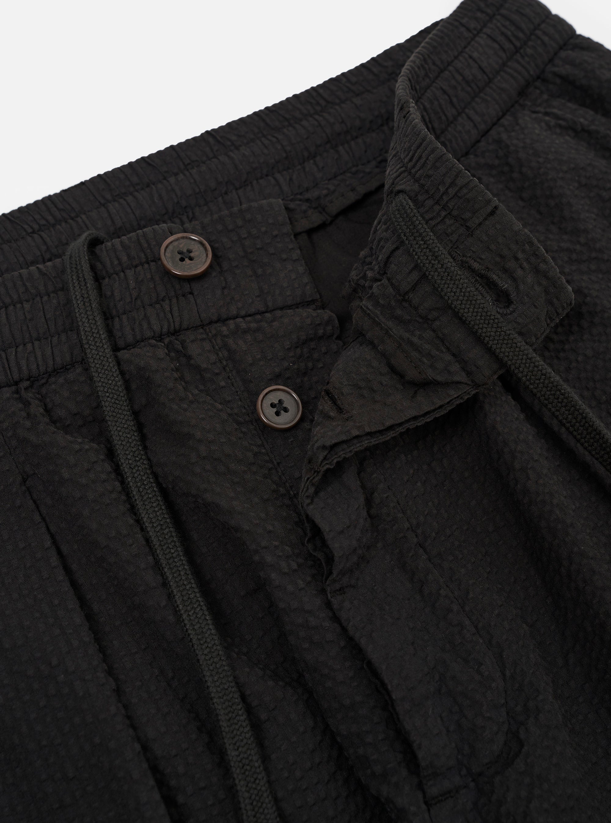 The Black Merida Seersucker Pleated Track Pant is shown from a rear three-quarter angle as a flat lay against a plain white background. The image highlights the elasticated waistband and the two rear pockets, consisting of one buttoned flap pocket and one button-jetted pocket. The black cotton fabric features a visible puckered seersucker texture and diagonal stitching details.