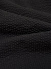 A close-up fabric swatch of the Black Merida Seersucker Pleated Track Pant highlights the solid black color and the distinct puckered texture of the seersucker fabric. The material fills the frame, providing a detailed view of the textile.