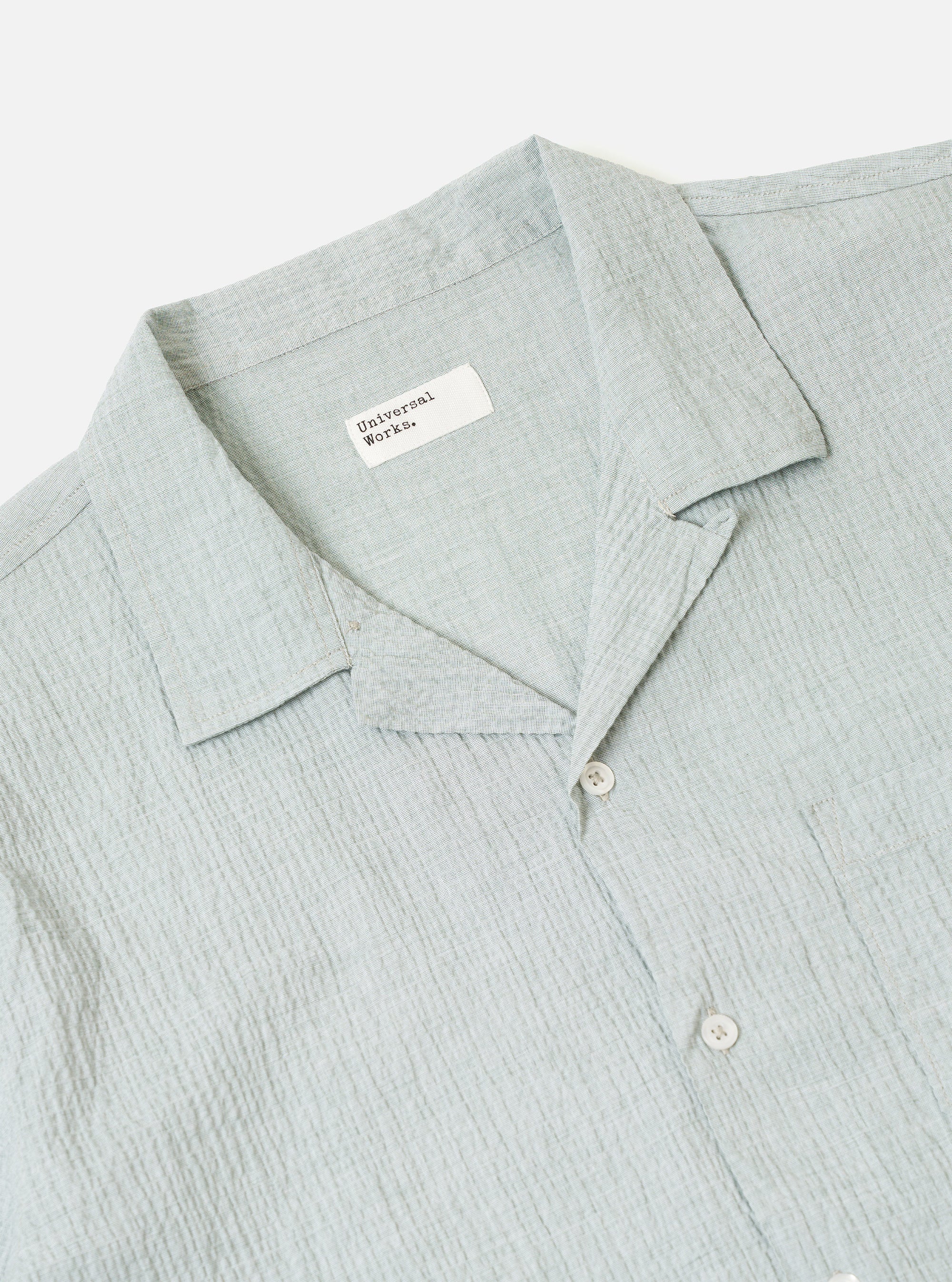 The Green Seersucker Road Shirt is worn by a model against a plain, neutral background. The shirt features a relaxed camp collar, a button-front closure, and a single chest pocket. The short-sleeve garment is made from a green cotton fabric with a distinct, puckered seersucker texture. The model is shown from the front, wearing the shirt untucked.