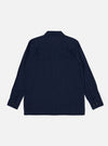 The Indigo Recycled Herringbone Attendant Shirt is worn by a model against a plain, light grey background. The model is shown from a front-side angle, wearing the shirt buttoned up. The image highlights the shirt's loose fit, one-piece flat collar, and two large button-flap patch pockets on the chest. The indigo-dyed fabric features a distinct herringbone weave texture.