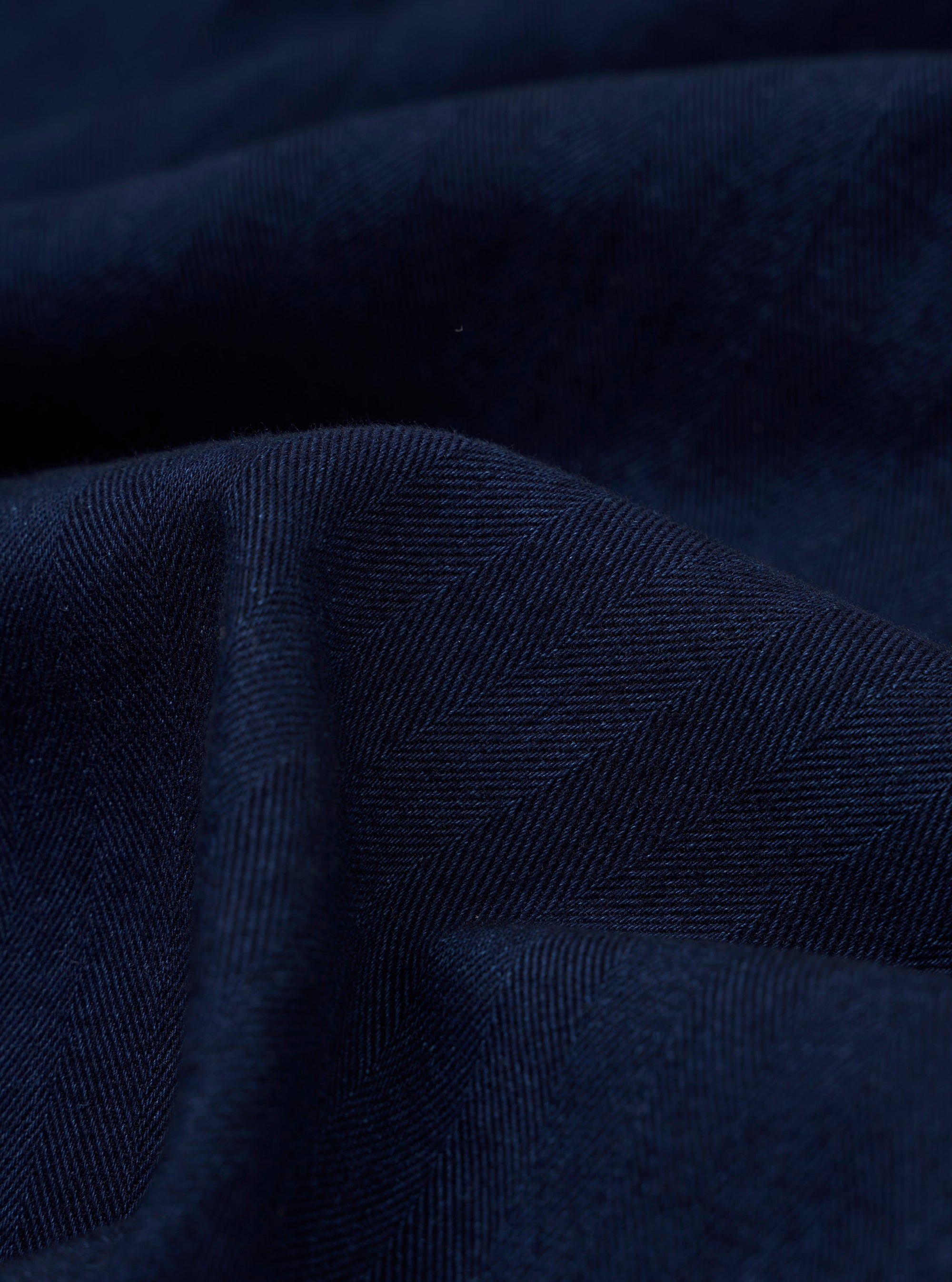 A close-up swatch image of the Indigo Recycled Herringbone Attendant Shirt fabric displays a deep indigo blue color with a distinct herringbone weave pattern. The texture of the recycled cotton fabric is prominently visible.