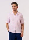 A model is wearing the Pink Lily Stripe Road Shirt, which features a camp collar, short sleeves, and a pink and white vertical stripe pattern. The model is shown from the waist up against a neutral, light-colored background. The shirt is unbuttoned over a white top, highlighting its relaxed fit and lightweight cotton fabric.