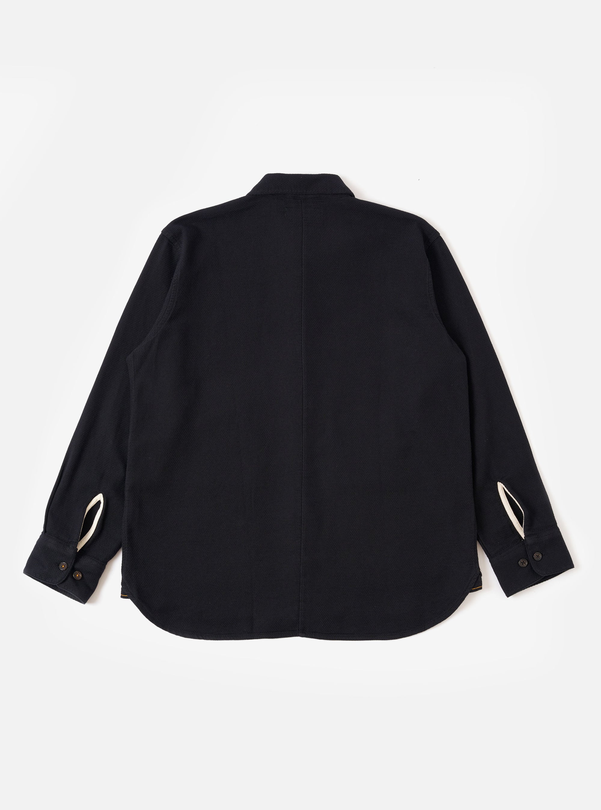 The Black Kendal Cotton Travail Overshirt is shown from the back as a flat lay against a plain white background. The image highlights the garment's loose, unstructured fit, curved hem, and a simple one-piece back panel. The black Kendal cotton fabric has a smooth, solid finish.