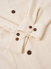 The Ecru Kendal Cotton Travail Overshirt is shown in a detail view as a flat lay against a plain white background. The image focuses on the lower corner of the garment, highlighting the curved shirt-like hem with a contrast stitch insert and the corner of a large patch pocket. The ecru-colored cotton fabric features a visible, textured open weave.