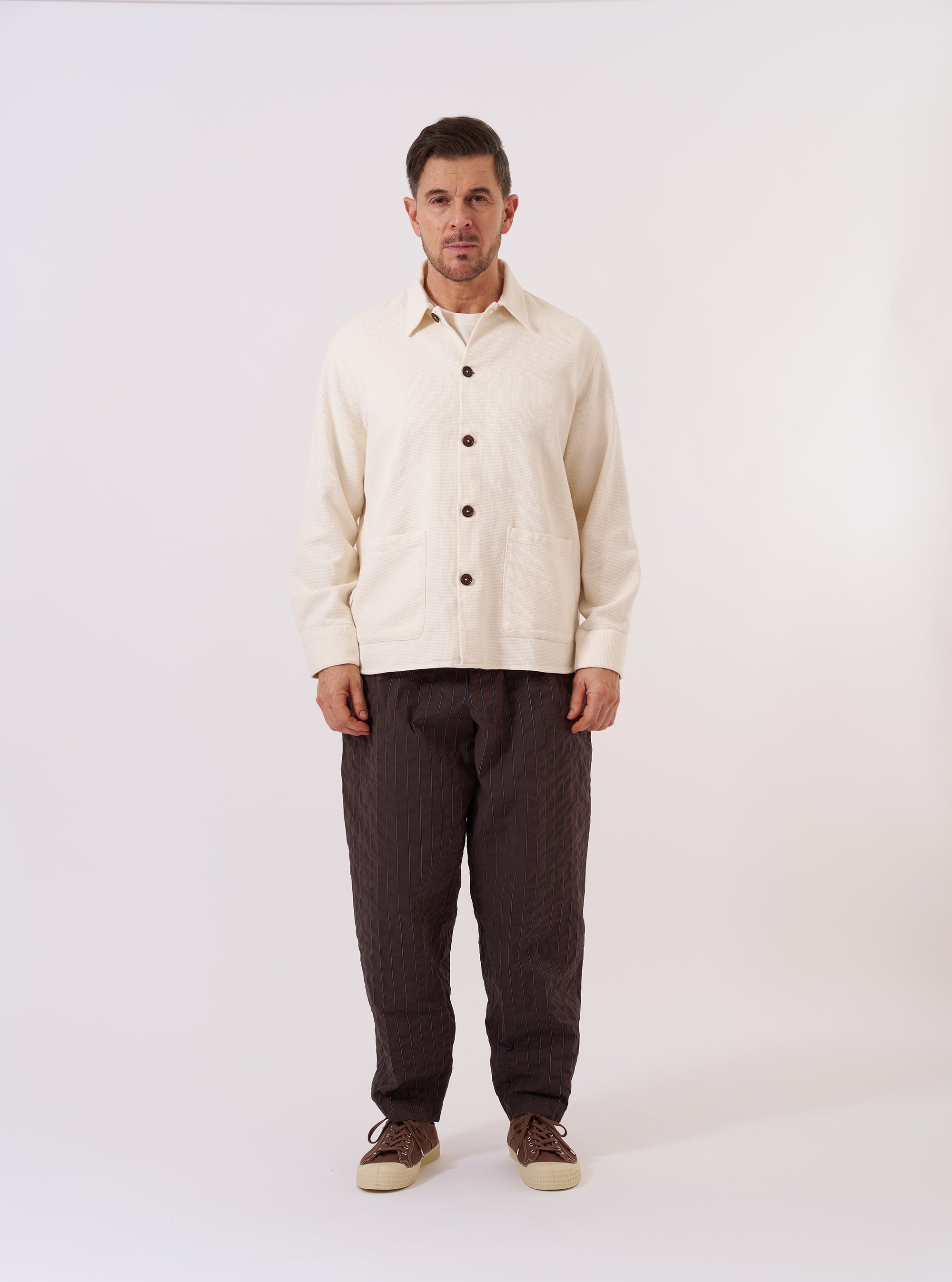 The Ecru Kendal Cotton Travail Overshirt is shown as a flat lay against a plain white background. The overshirt features a classic one-piece collar, a central button-down closure, and two large patch pockets at the waist. The textured ecru cotton fabric has a visible open weave, and the garment is finished with a curved hem and adjustable button cuffs.