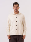 A model wears the Ecru Kendal Cotton Travail Overshirt against a plain, neutral background. The overshirt features a one-piece collar, a central button-down closure, and two large patch pockets at the waist. The garment is made from an off-white ecru cotton fabric and has a loose, unstructured fit.