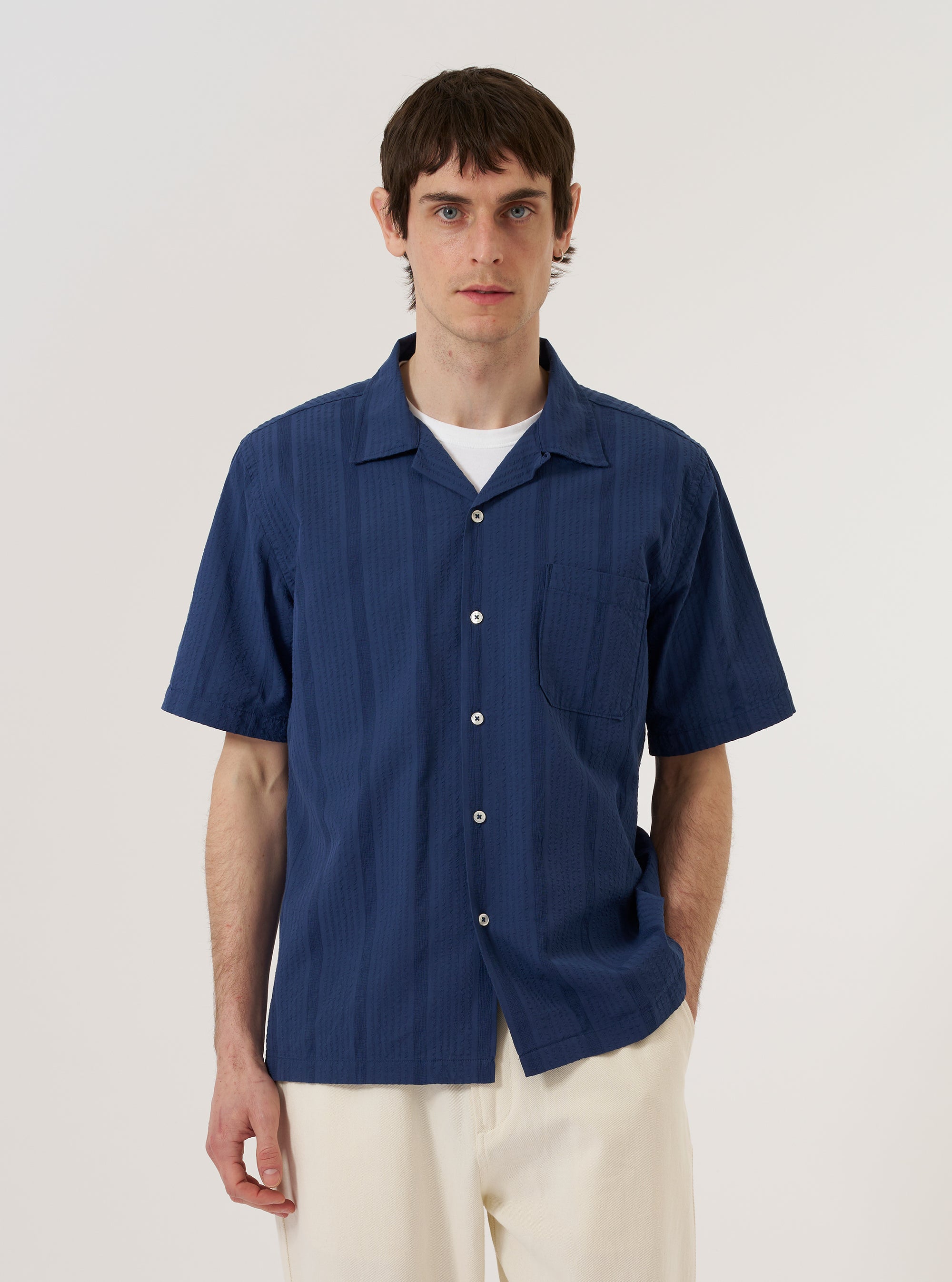 The Blue Maui Cotton Road Shirt is worn by a model against a plain white background. The shirt features a vertical striped pattern in blue and navy, a camp collar, and a single chest pocket on the left side. The model is shown from the chest up, with the shirt buttoned.