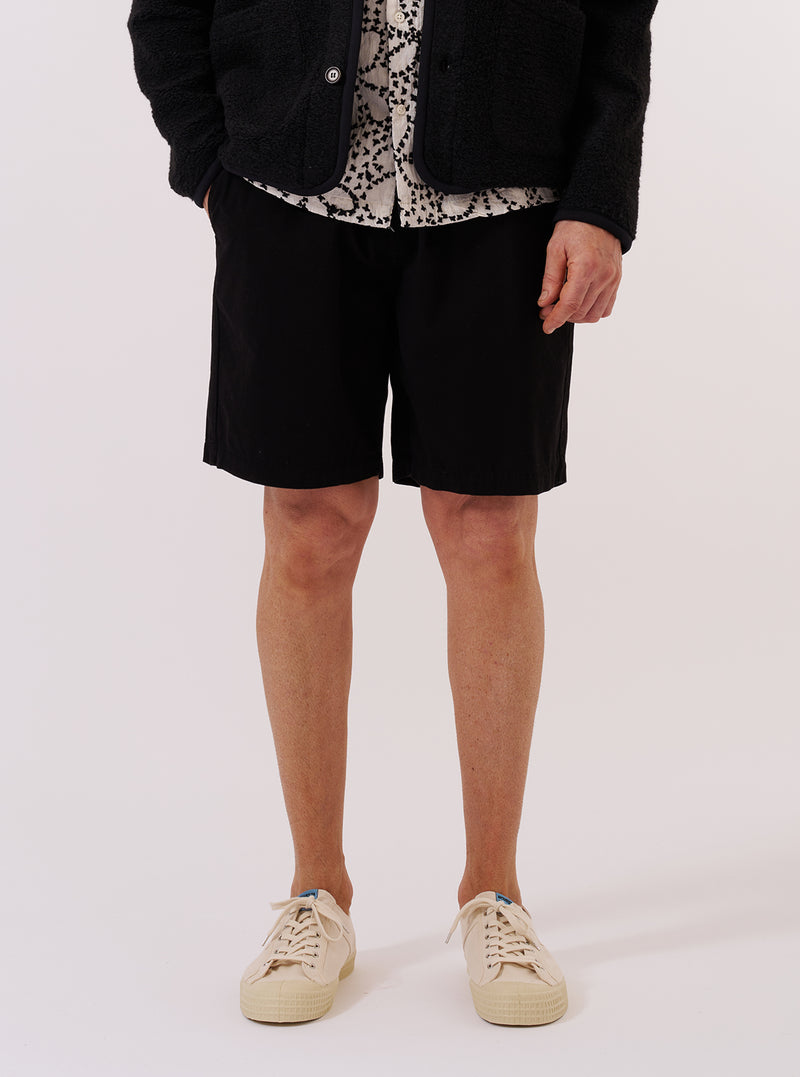 A model wears the Black Kelly Cotton Simple Short against a plain white background. The shorts feature a relaxed fit, an elasticated waistband, and side slant pockets. The black cotton fabric has a clean, smooth finish. The model is shown from the waist down, wearing the shorts with a white t-shirt and black shoes.